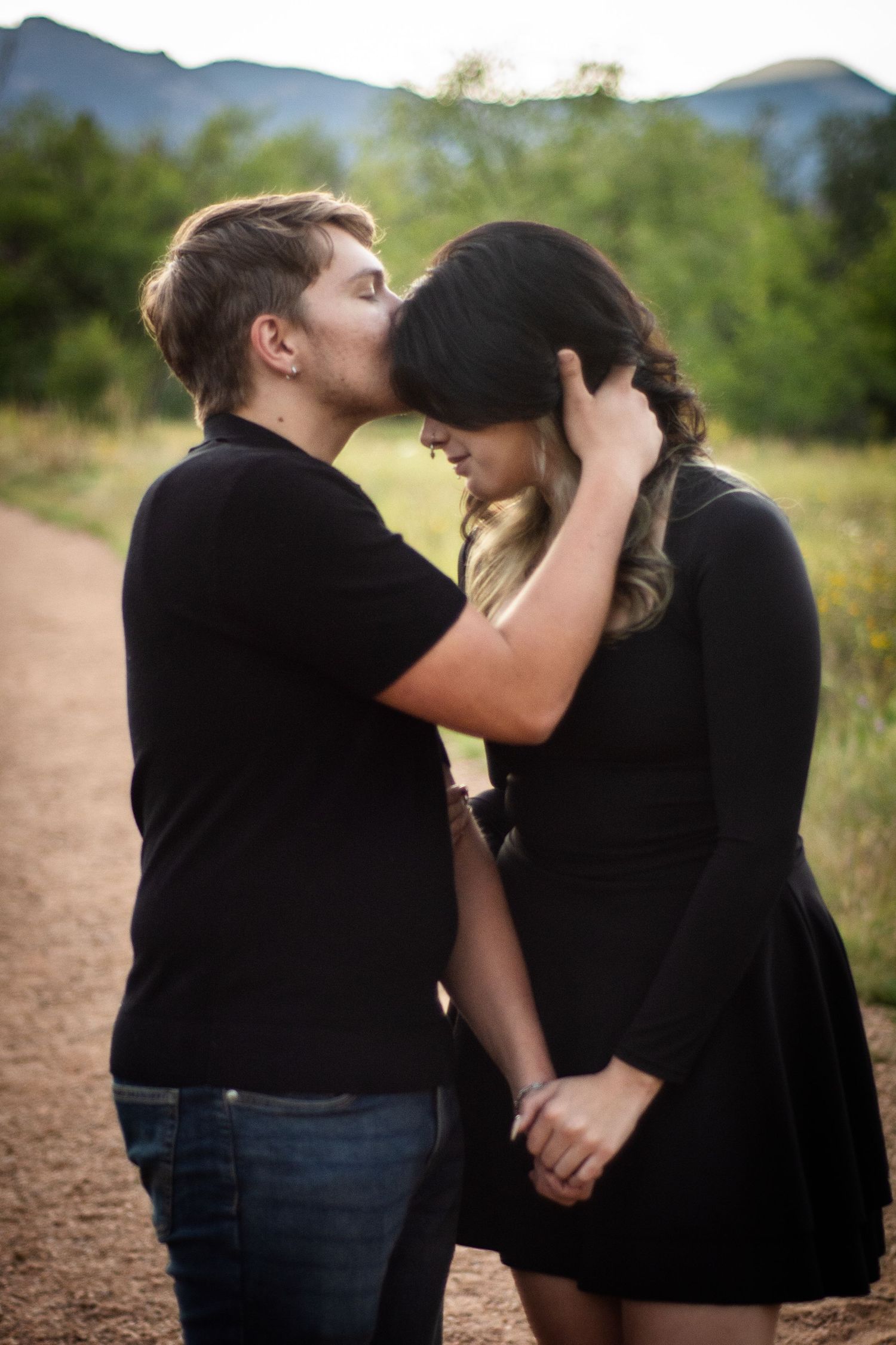 Couple sharing a tender kiss on a dirt path with mountains visible in the background.