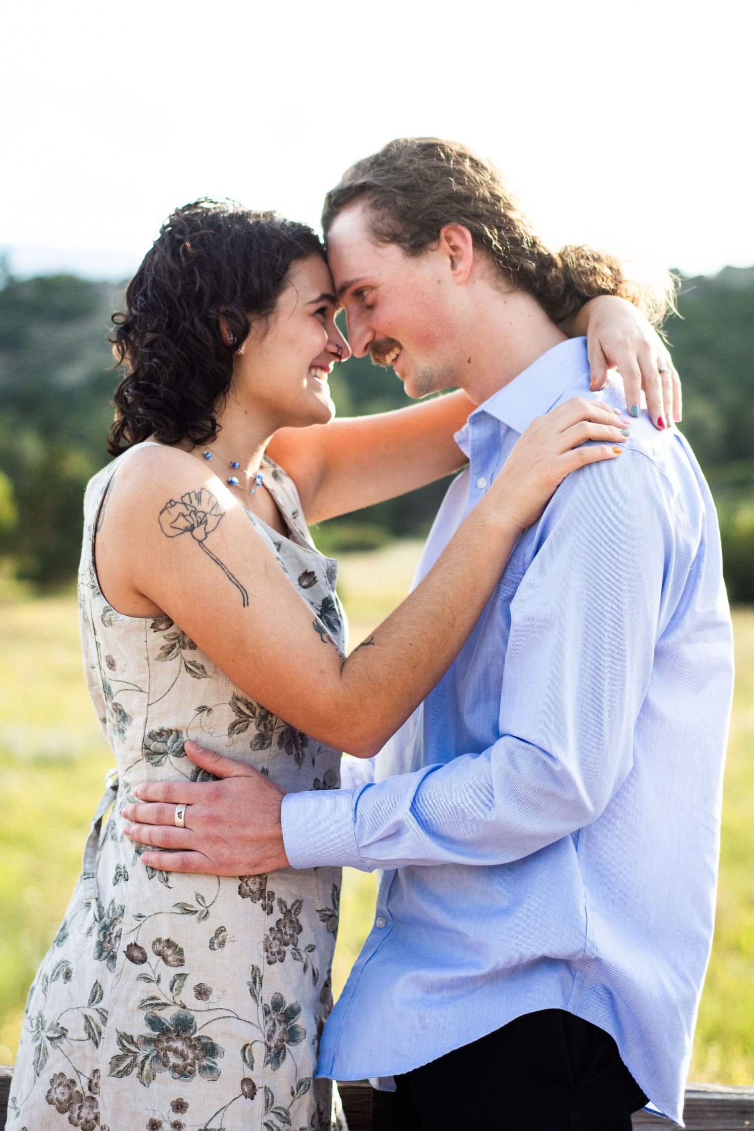 Smiling couple embracing forehead-to-forehead in a sunny outdoor field, woman with floral tattoo.