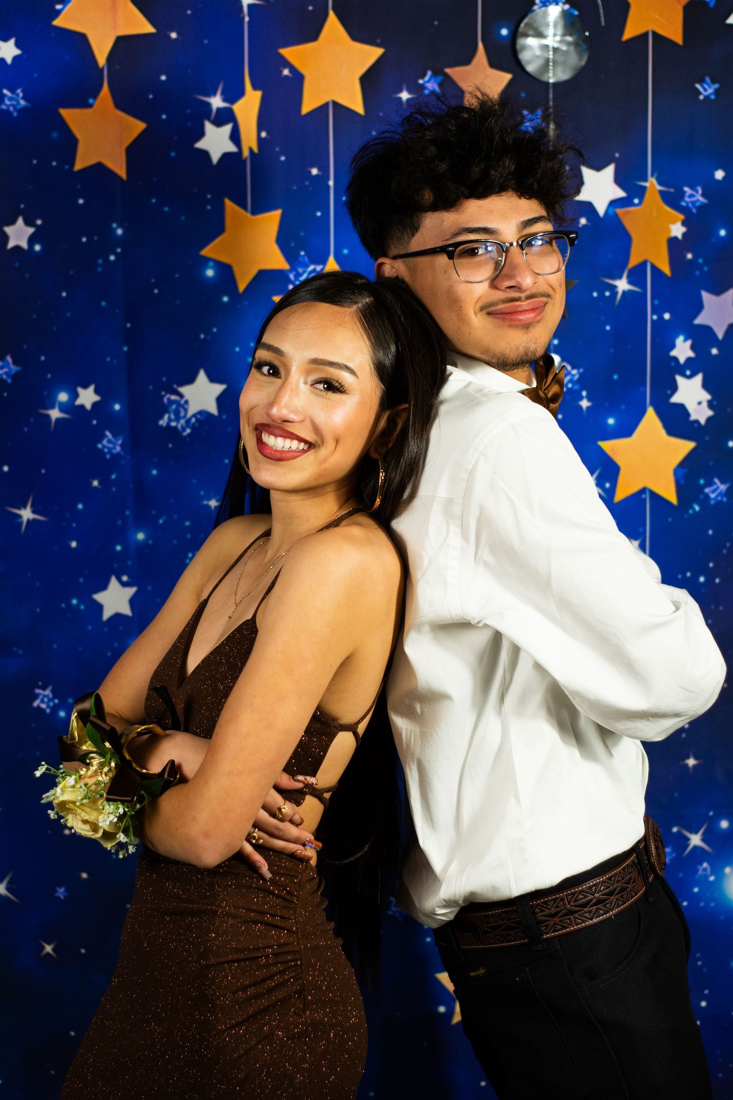 A couple poses back-to-back in formal prom attire against a blue starry backdrop.