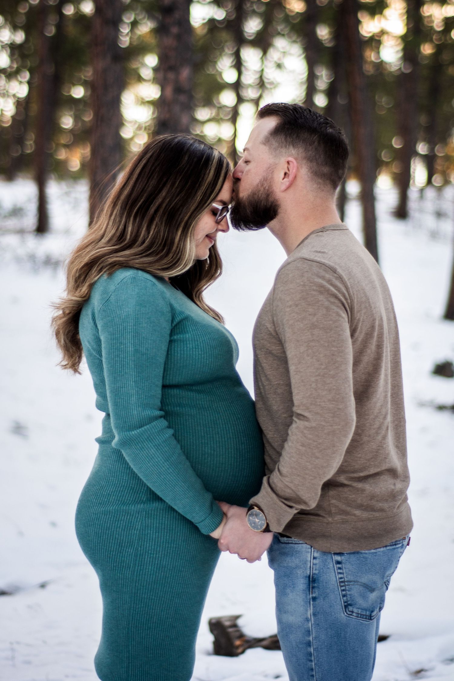 Couple sharing a kiss during a winter maternity photo shoot in a snowy forest, woman in teal dress showing baby bump.