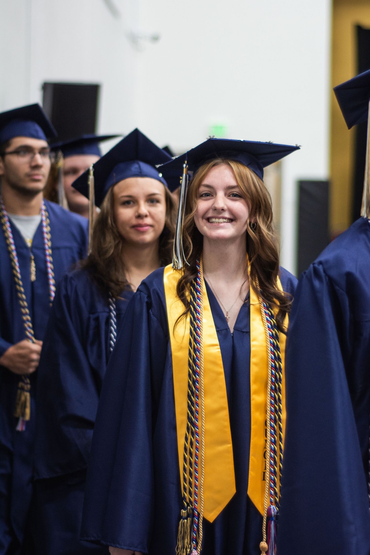Smiling female graduates in navy blue caps and gowns with gold honor cords at graduation ceremony.