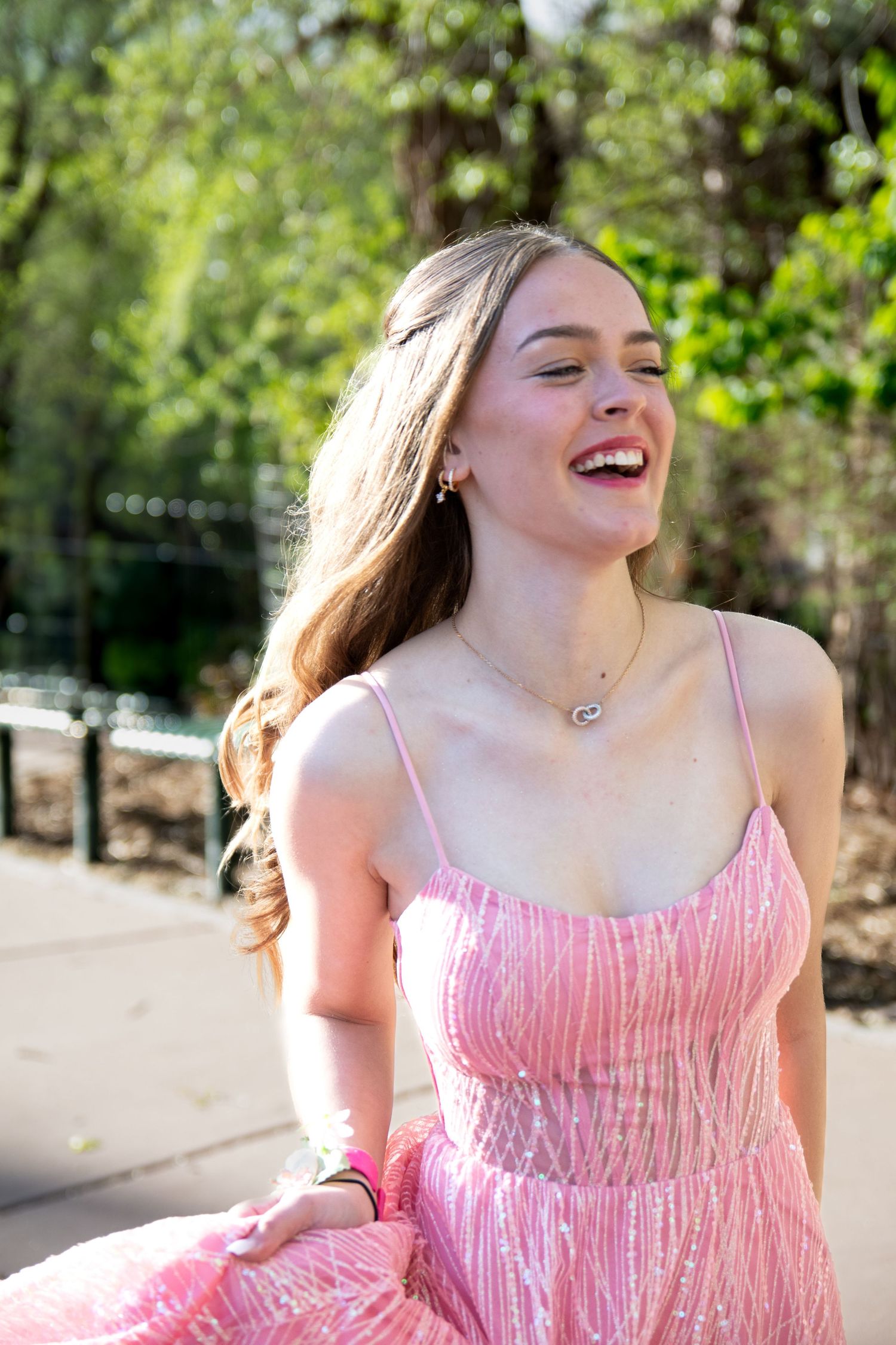 Young woman in pink sequin dress laughing outdoors in sunlight, with long hair and delicate necklace.