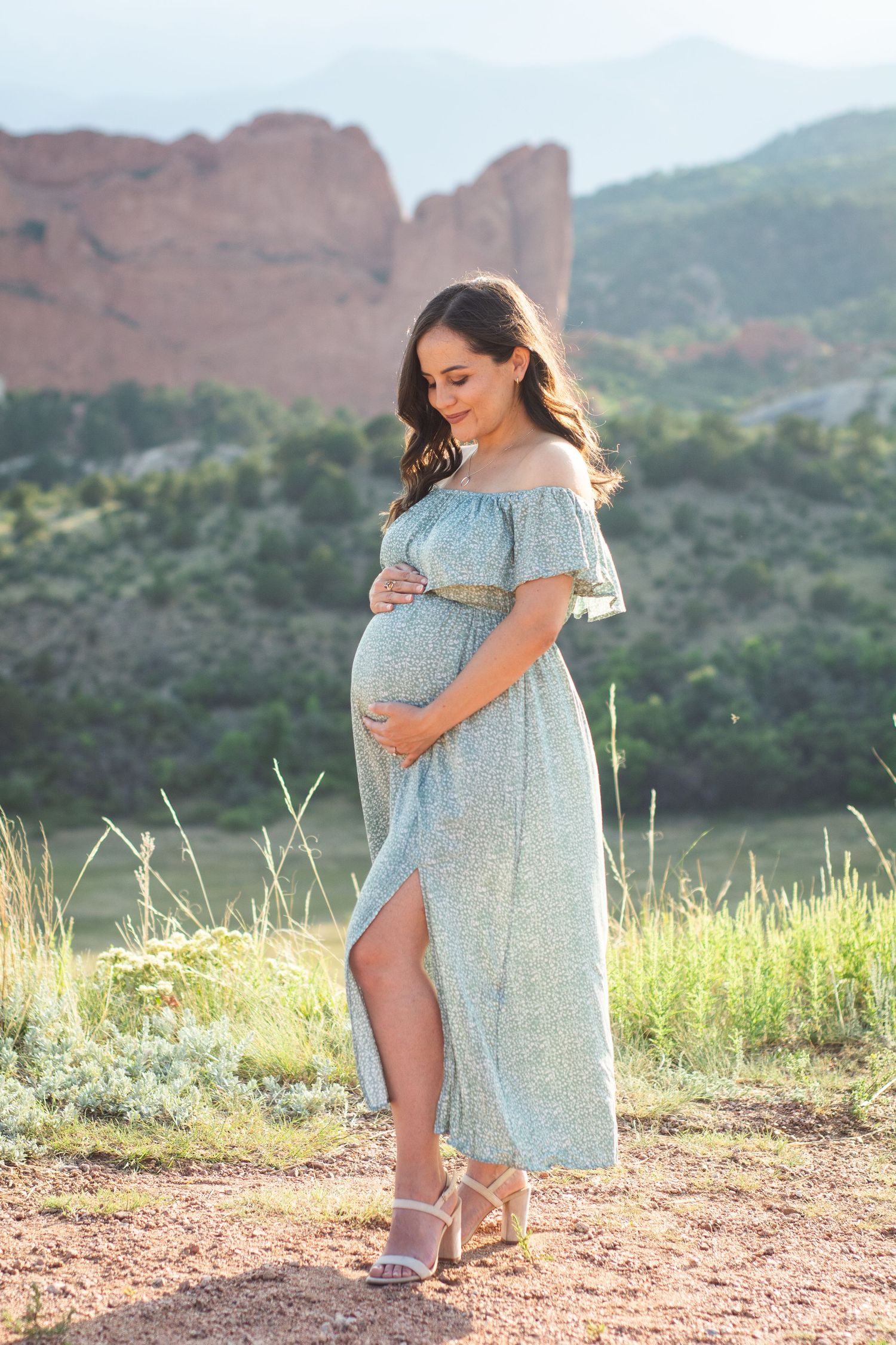 Pregnant woman in a light blue off-shoulder maxi dress cradling her baby bump outdoors with red rock formations behind her.