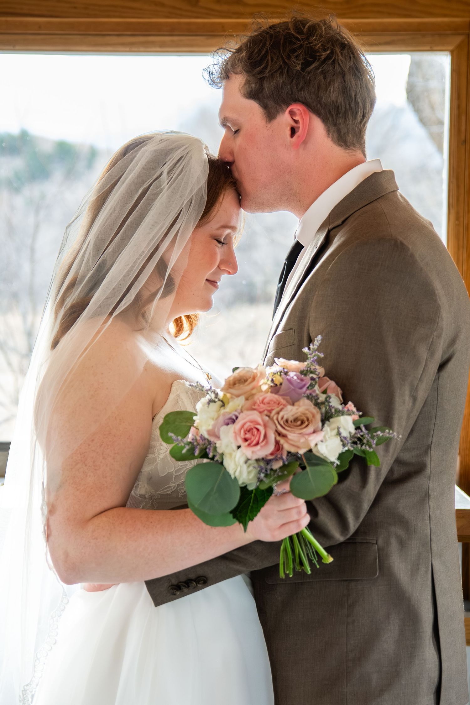 Bride and groom share tender moment, she holds pastel rose bouquet, he kisses her forehead, wearing veil and gray suit.