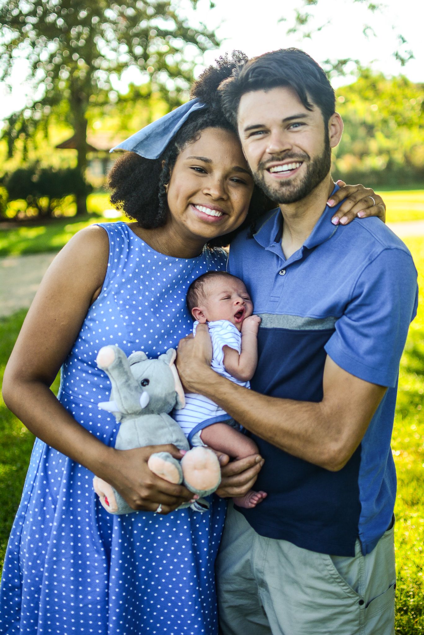 A smiling interracial couple poses outdoors holding their newborn baby and a stuffed elephant toy in a sunny park.
