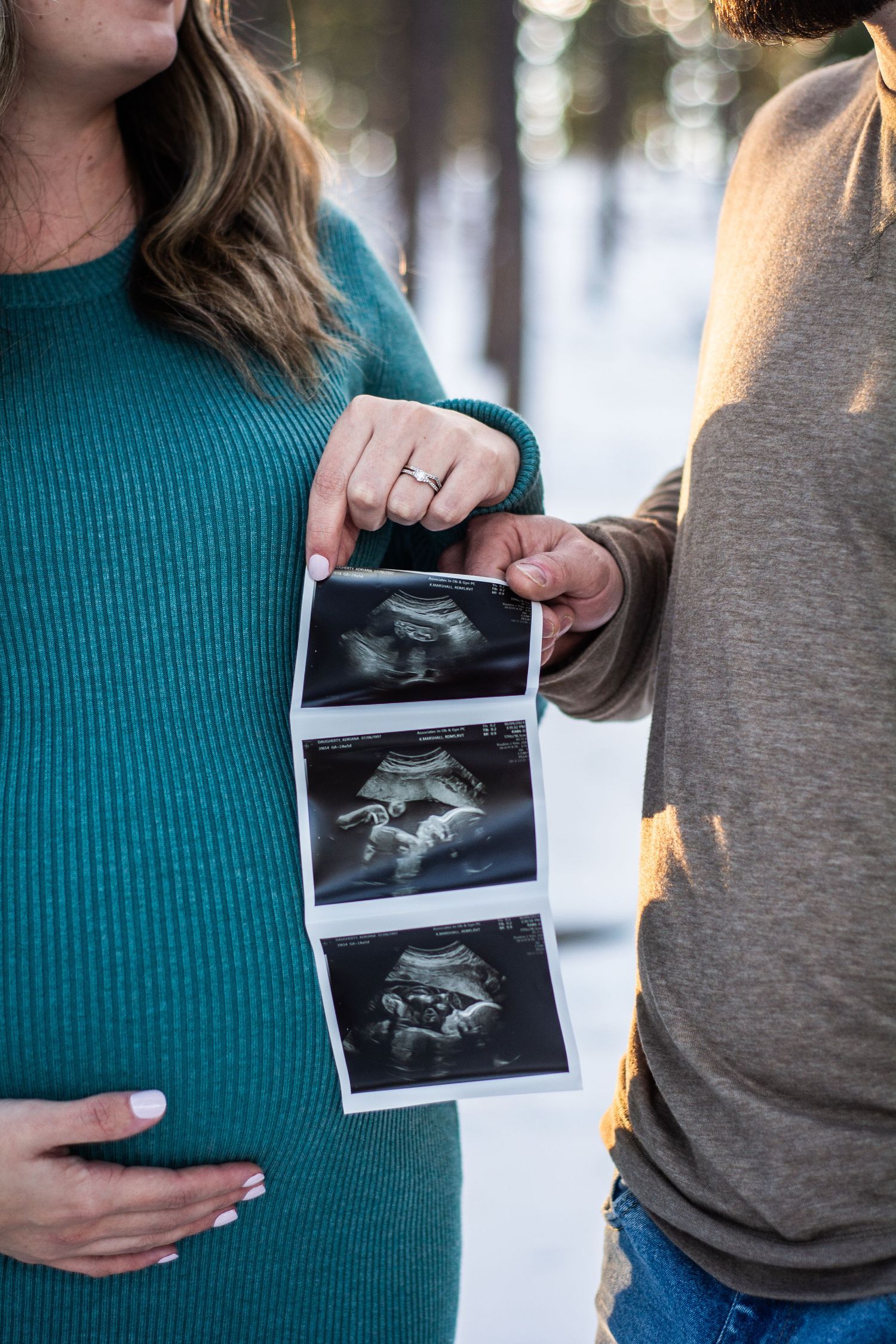 Pregnant couple holding ultrasound photos outdoors in snowy winter forest, woman in teal sweater cradling baby bump.