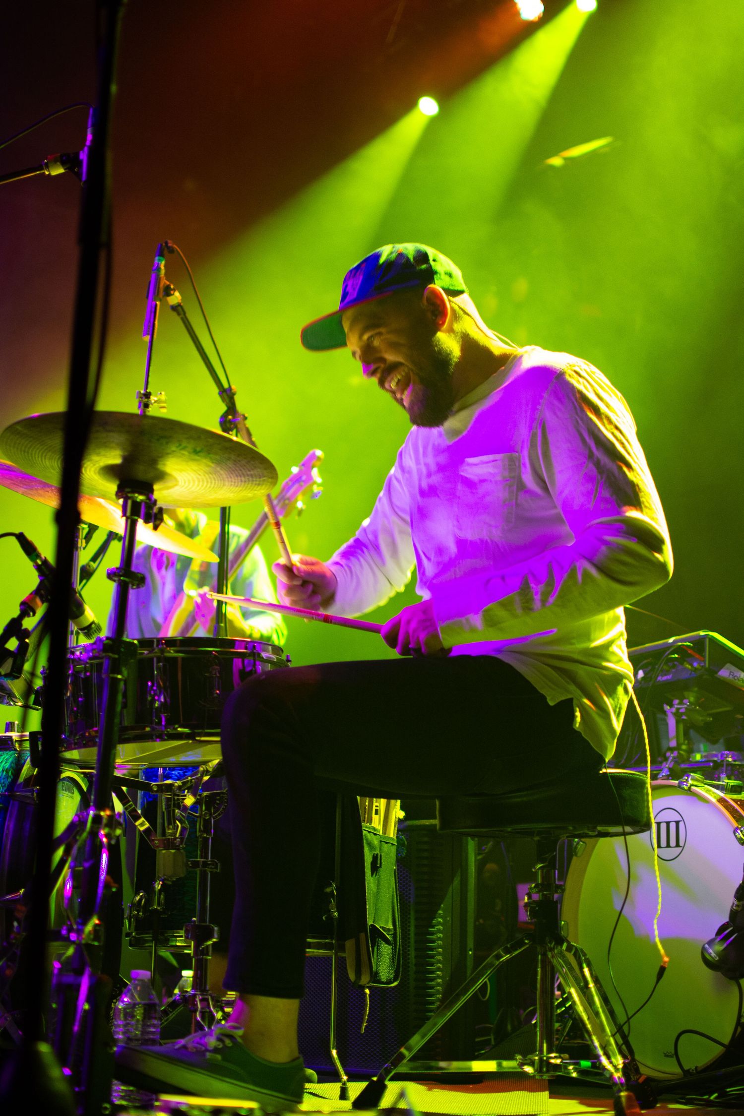 An energetic drummer performs on stage under vibrant green and purple stage lighting.
