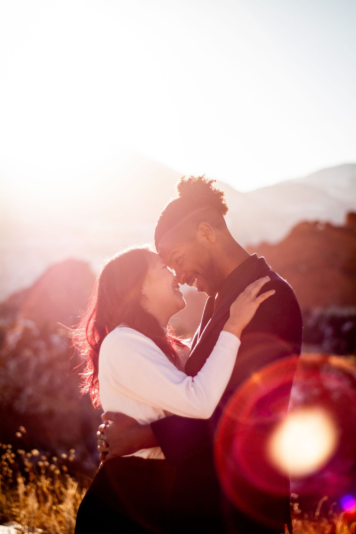 Romantic couple touching foreheads at golden sunset with mountain backdrop and lens flare.