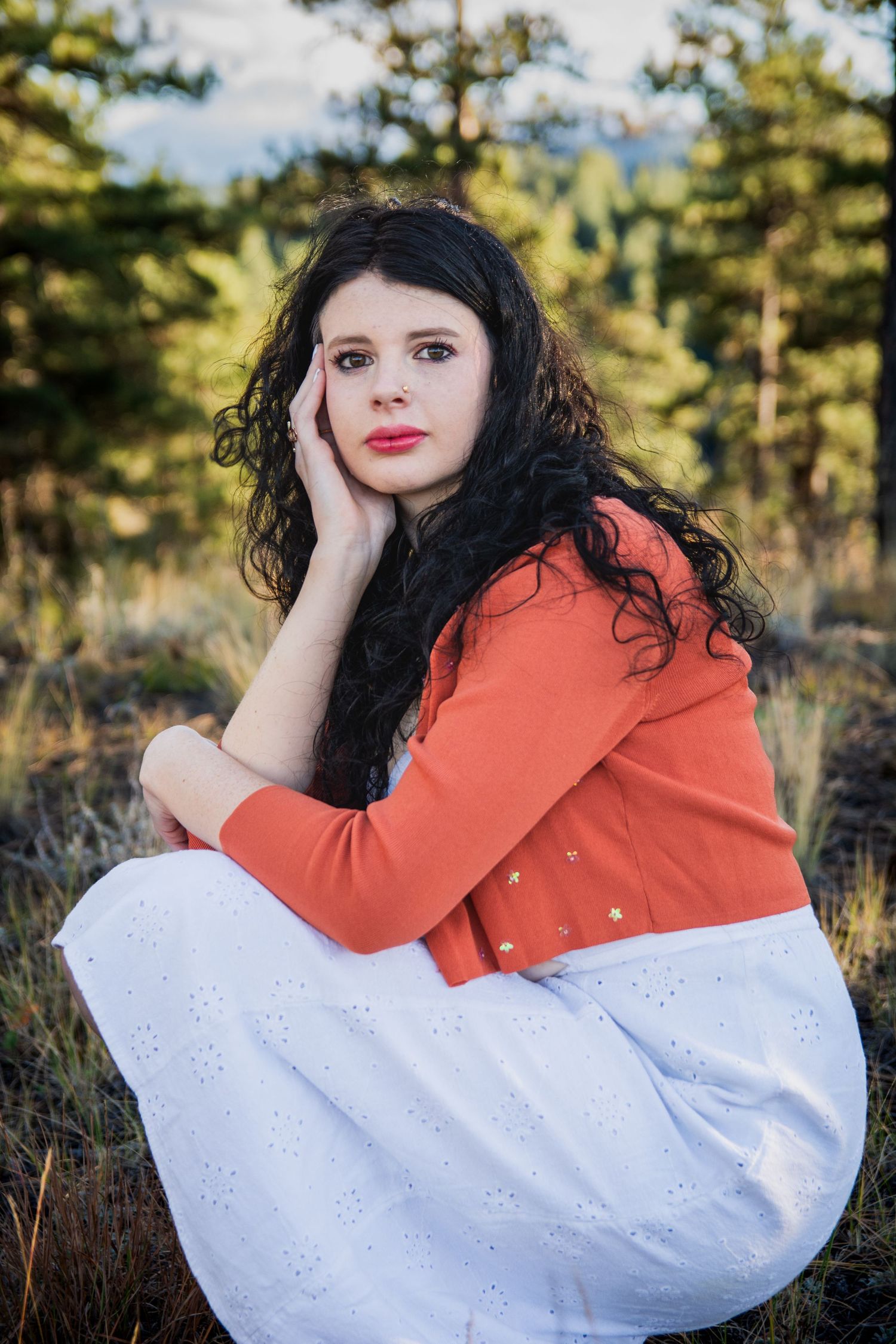 A young woman with dark curly hair in an orange cardigan and white skirt sits outdoors among pine trees.