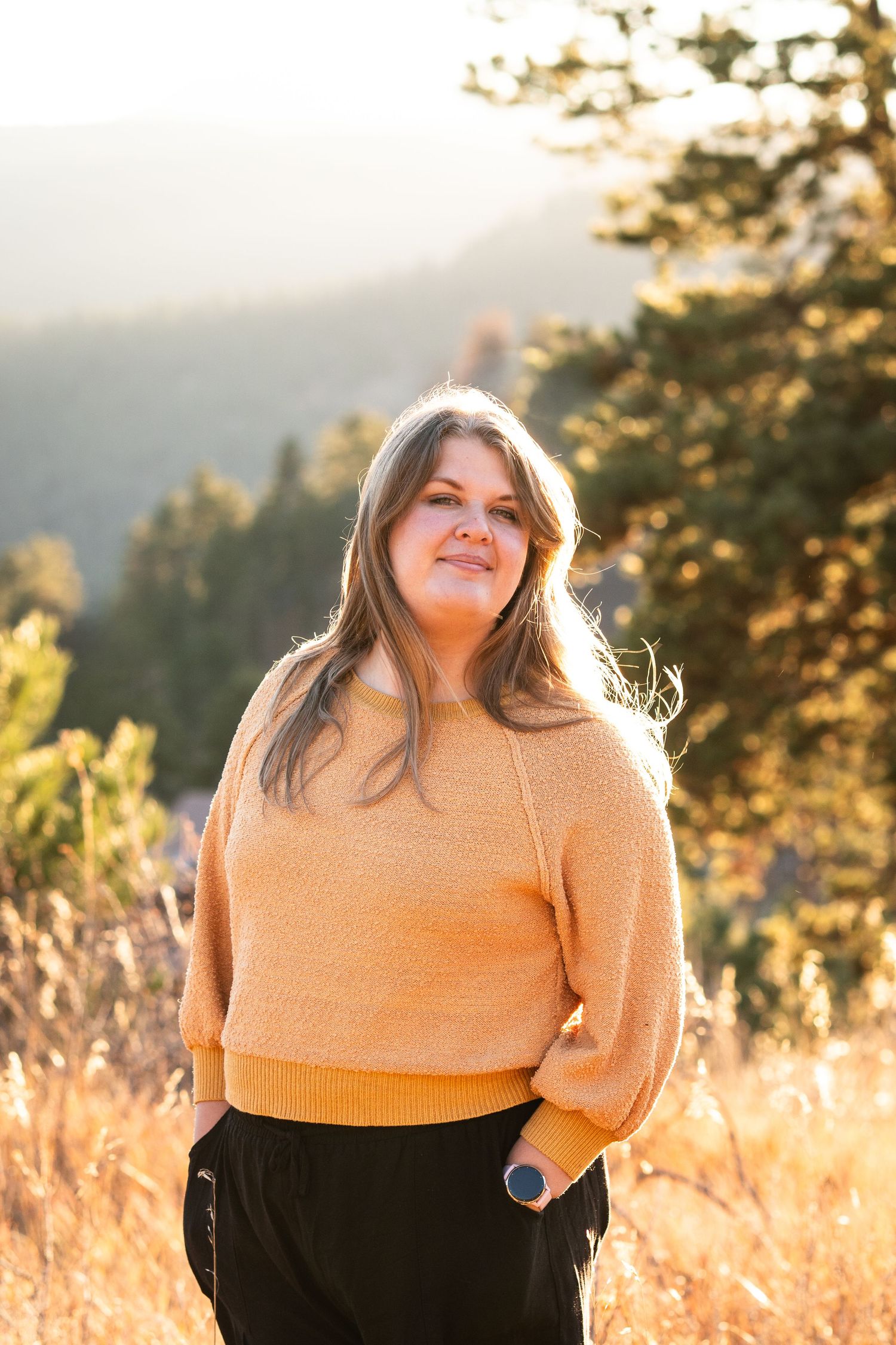 Young woman in orange sweater smiling outdoors in golden sunlight with mountain landscape background.