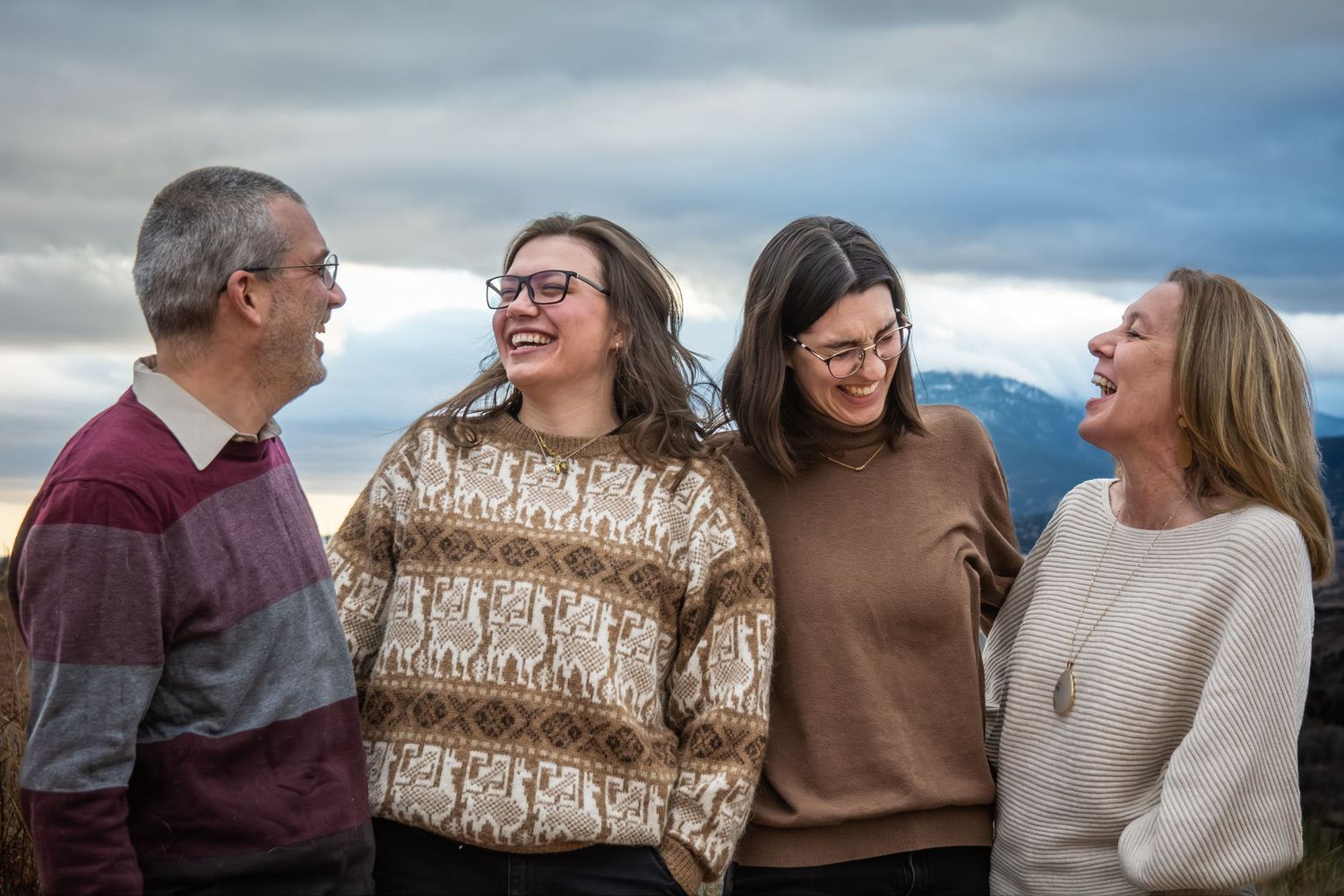 Family of four laughing together outdoors with mountains and cloudy sky in the background.