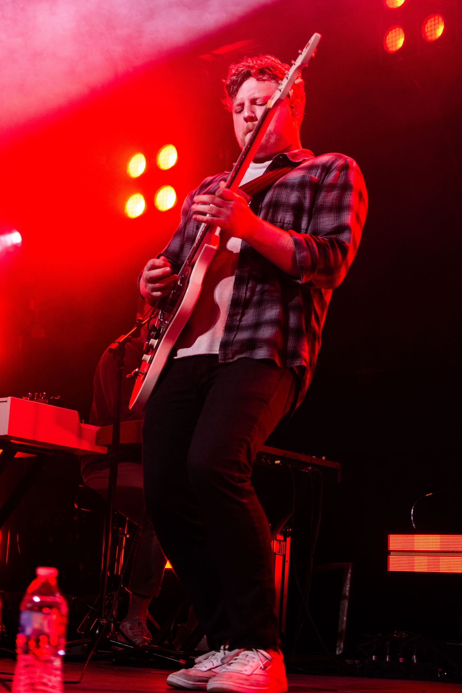 A guitarist performs on stage bathed in dramatic red concert lighting.