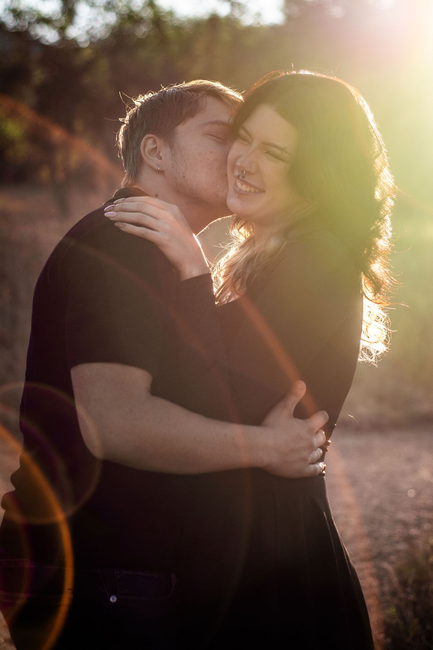 Man kissing smiling woman on cheek while embracing outdoors in warm golden backlight.