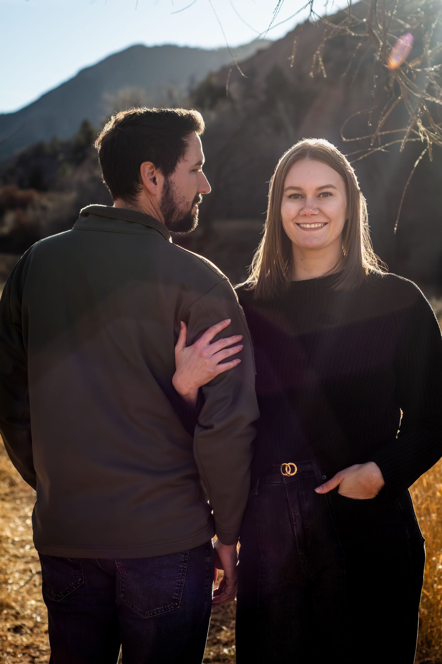 Couple smiling together outdoors with mountain backdrop. The woman is resting her hand around the man's arm as he looks at her.