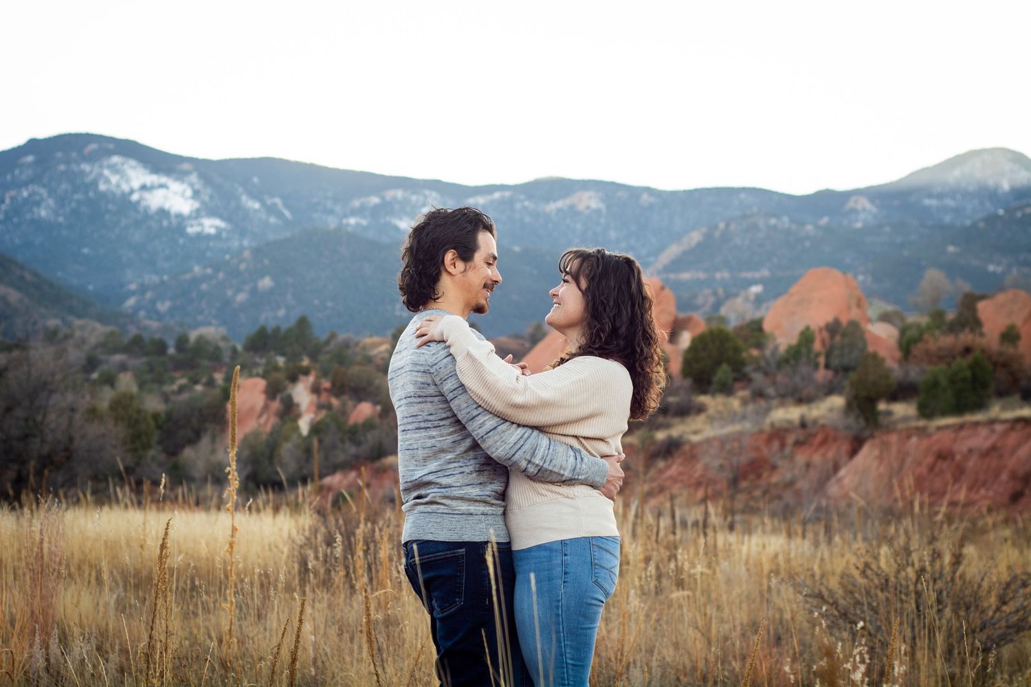 Couple embracing in golden field with red rock formations and mountains behind them.