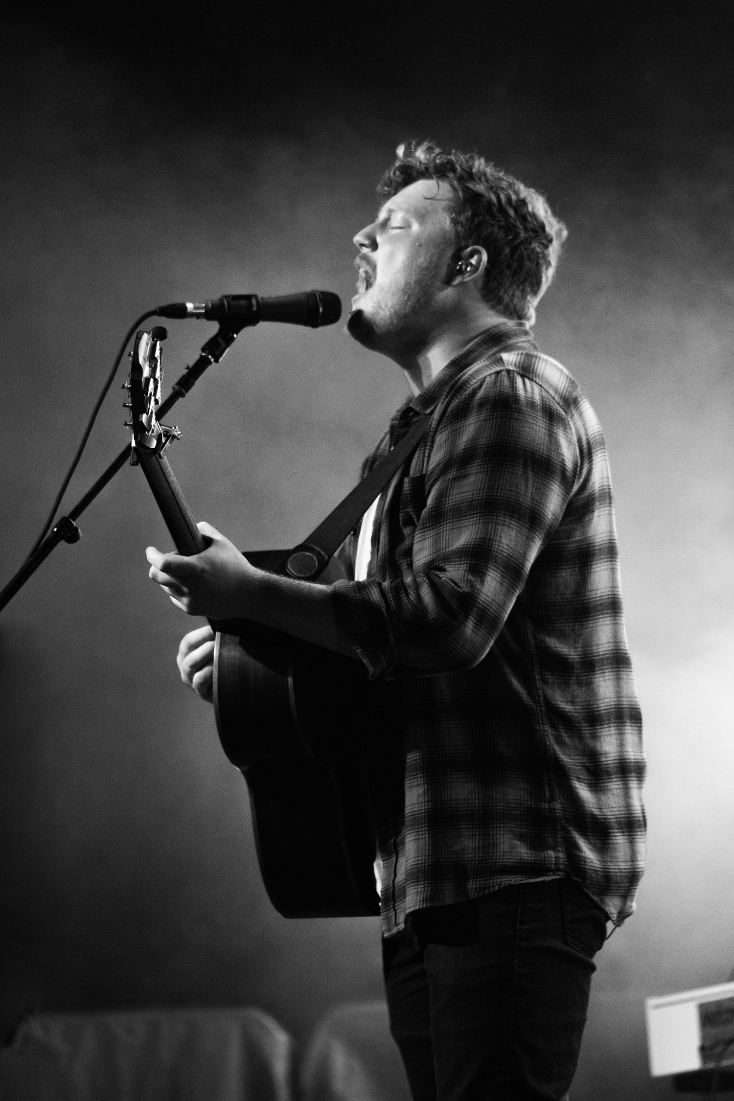 Black and white photo of a male musician playing acoustic guitar and singing into a microphone on stage.