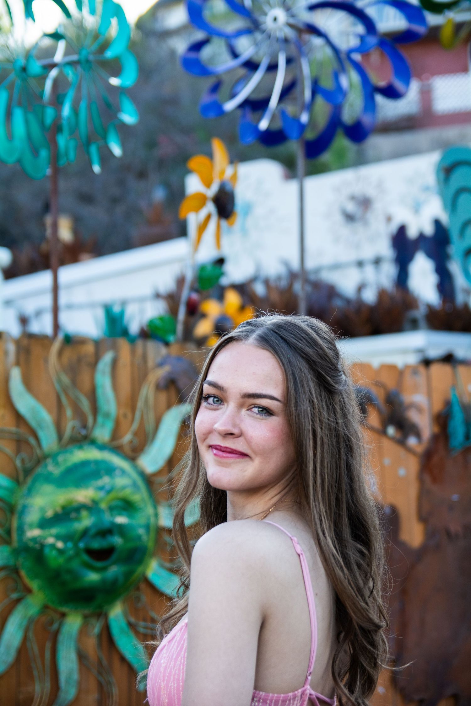 Young woman with long brown hair smiles over shoulder near colorful metal garden art and pinwheels on wooden fence.