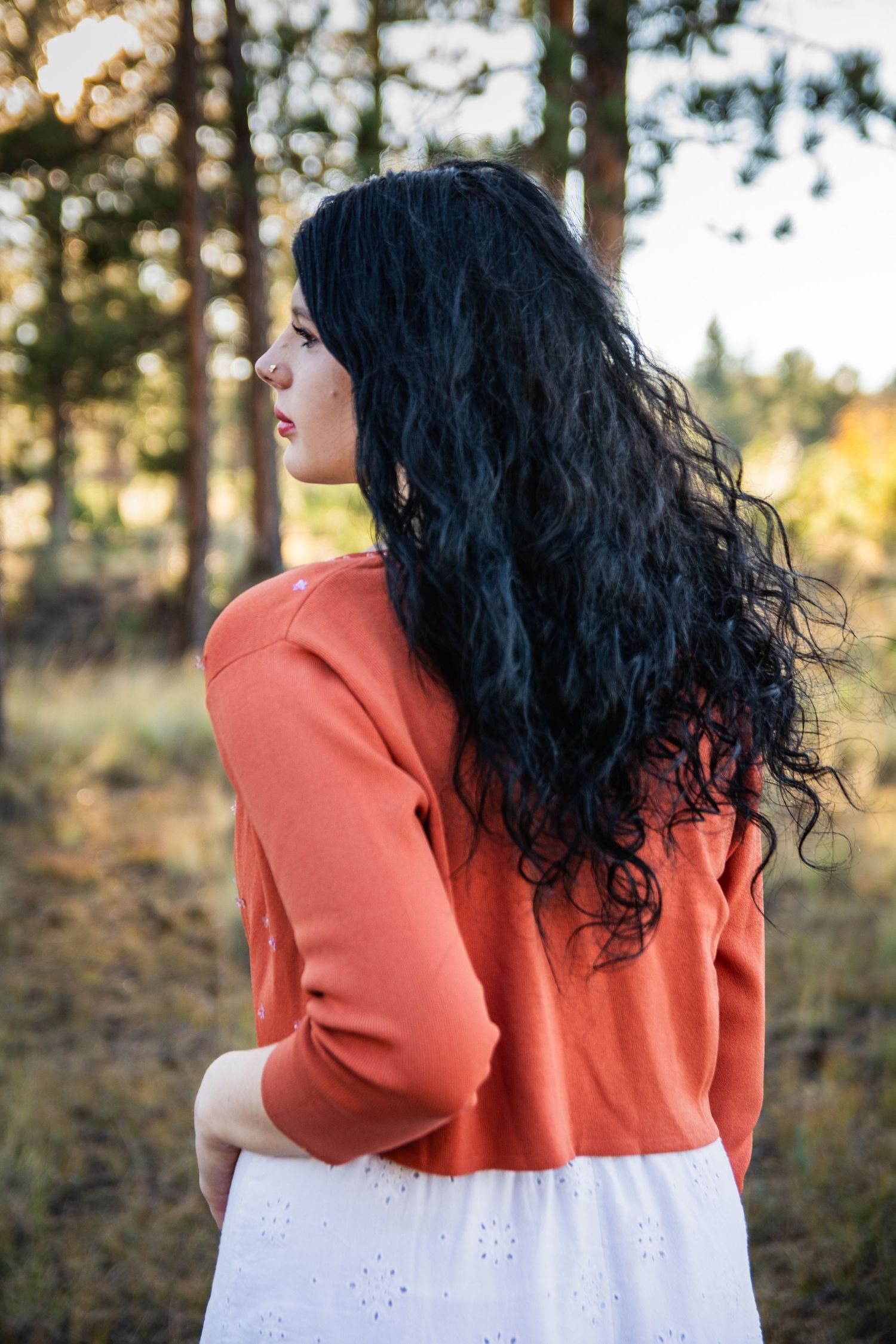 Woman with long black curly hair wearing an orange sweater and white skirt standing in a forest.
