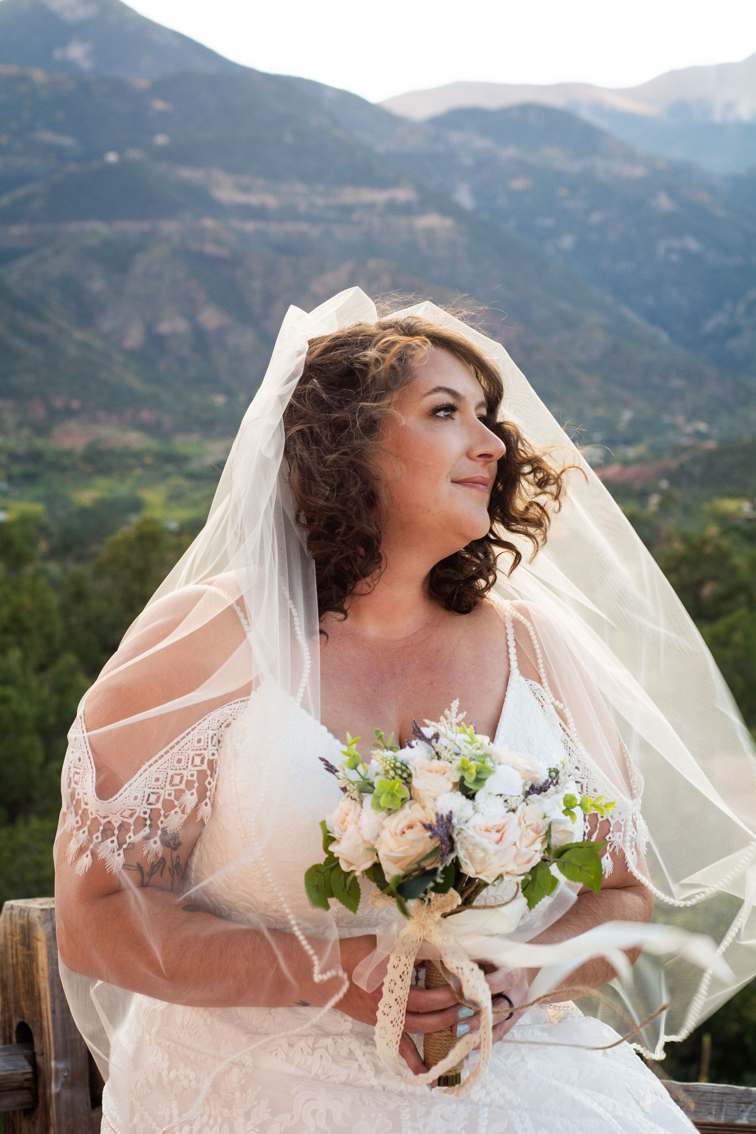Bride in lace wedding dress and veil holds bouquet with mountain landscape backdrop.