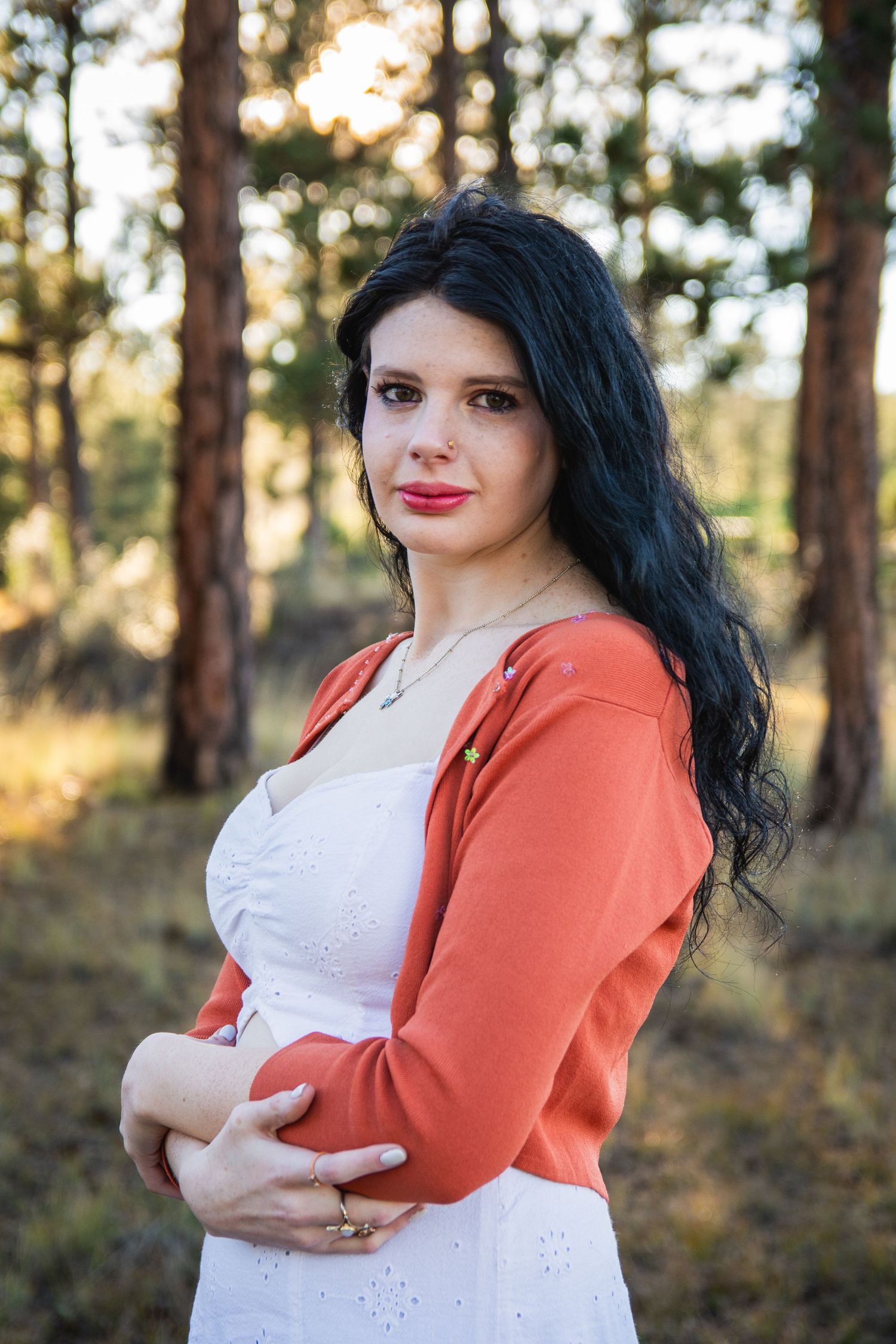 A young woman with long dark hair wearing a white dress and orange cardigan poses in a sunlit forest setting.