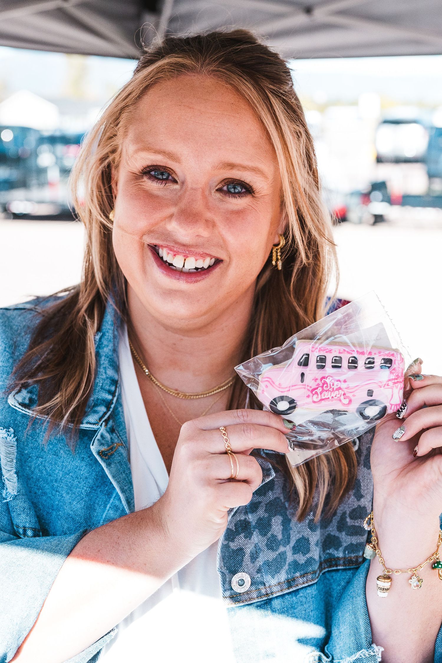 Smiling young woman with blonde hair holding a pink bus-shaped decorated cookie in a plastic bag at an outdoor event.
