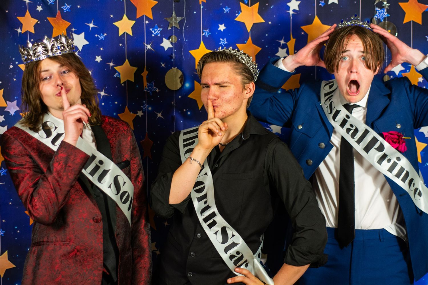 Three prom royalty winners wearing sashes pose humorously against a festive blue starry backdrop.