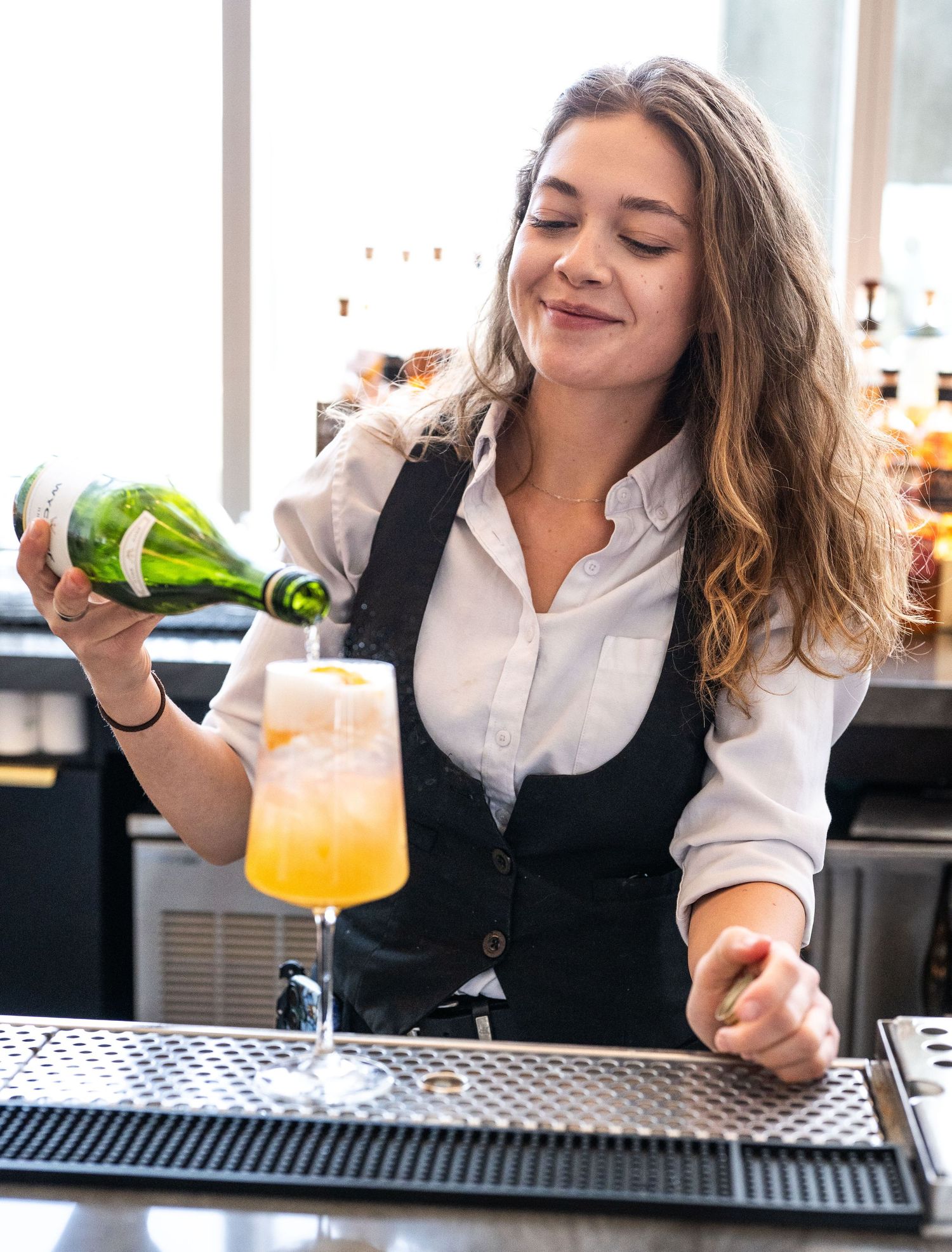 Female bartender pouring a green bottle into an orange cocktail in a wine glass at a bar.