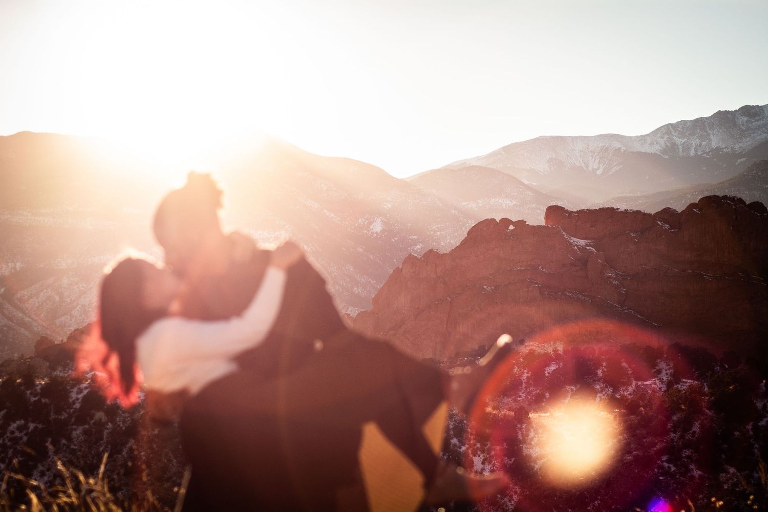 Couple sharing a kiss backlit by bright golden sunset with rocky mountain landscape behind.