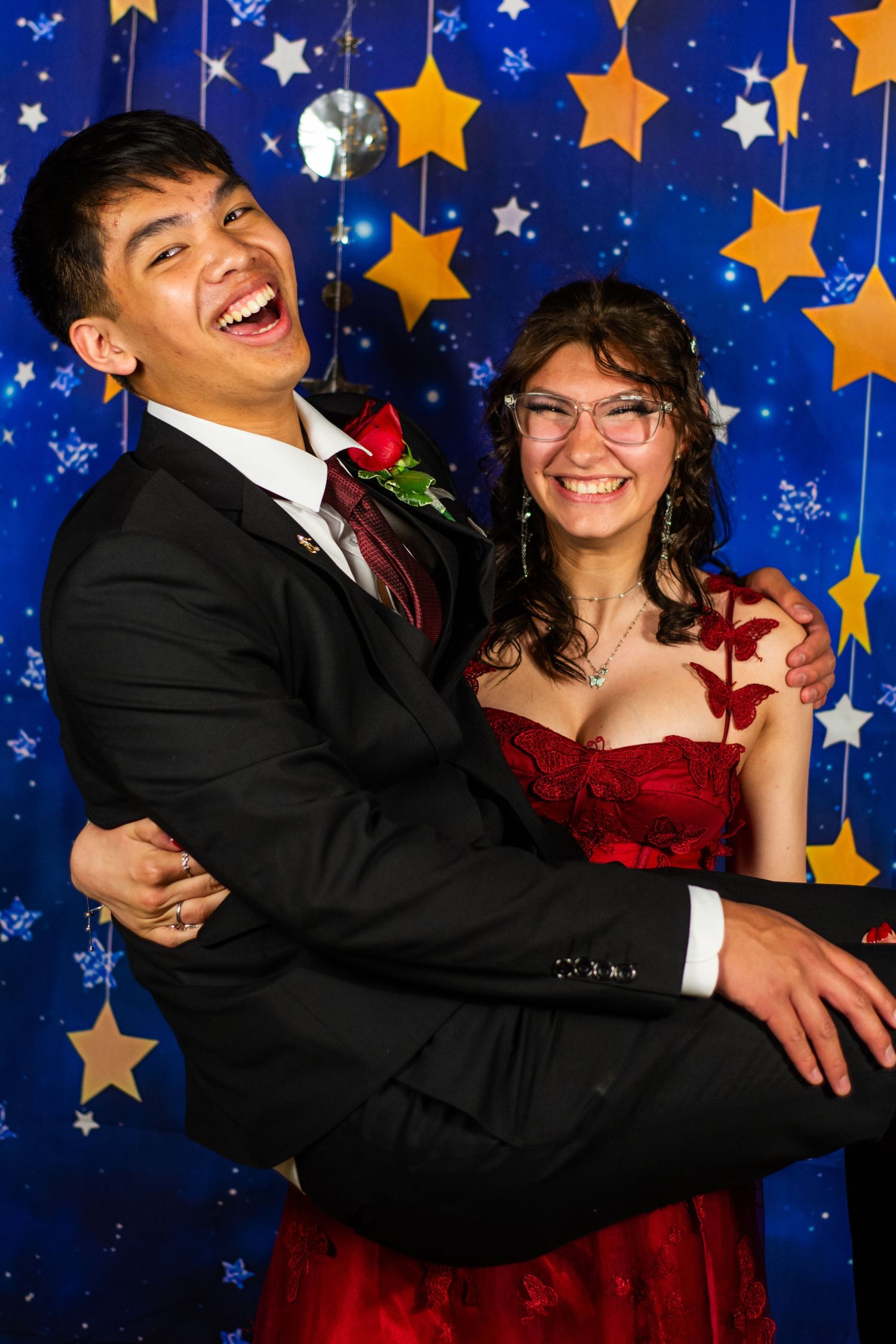 A laughing young couple poses playfully in formal attire in front of a star-themed prom backdrop.