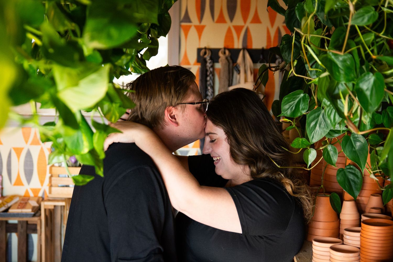 Couple sharing a romantic moment surrounded by lush green plants and terracotta pots in a cozy indoor setting.