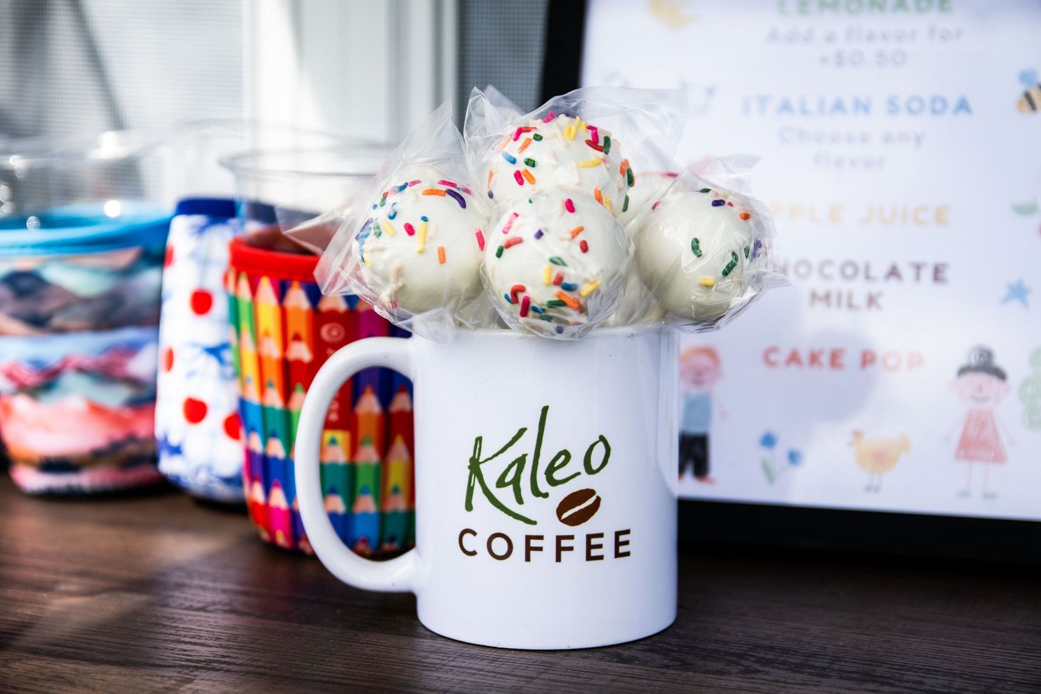 Kaleo Coffee mug filled with colorful sprinkle cake pops on a wooden table with crayons and a menu board in the background.