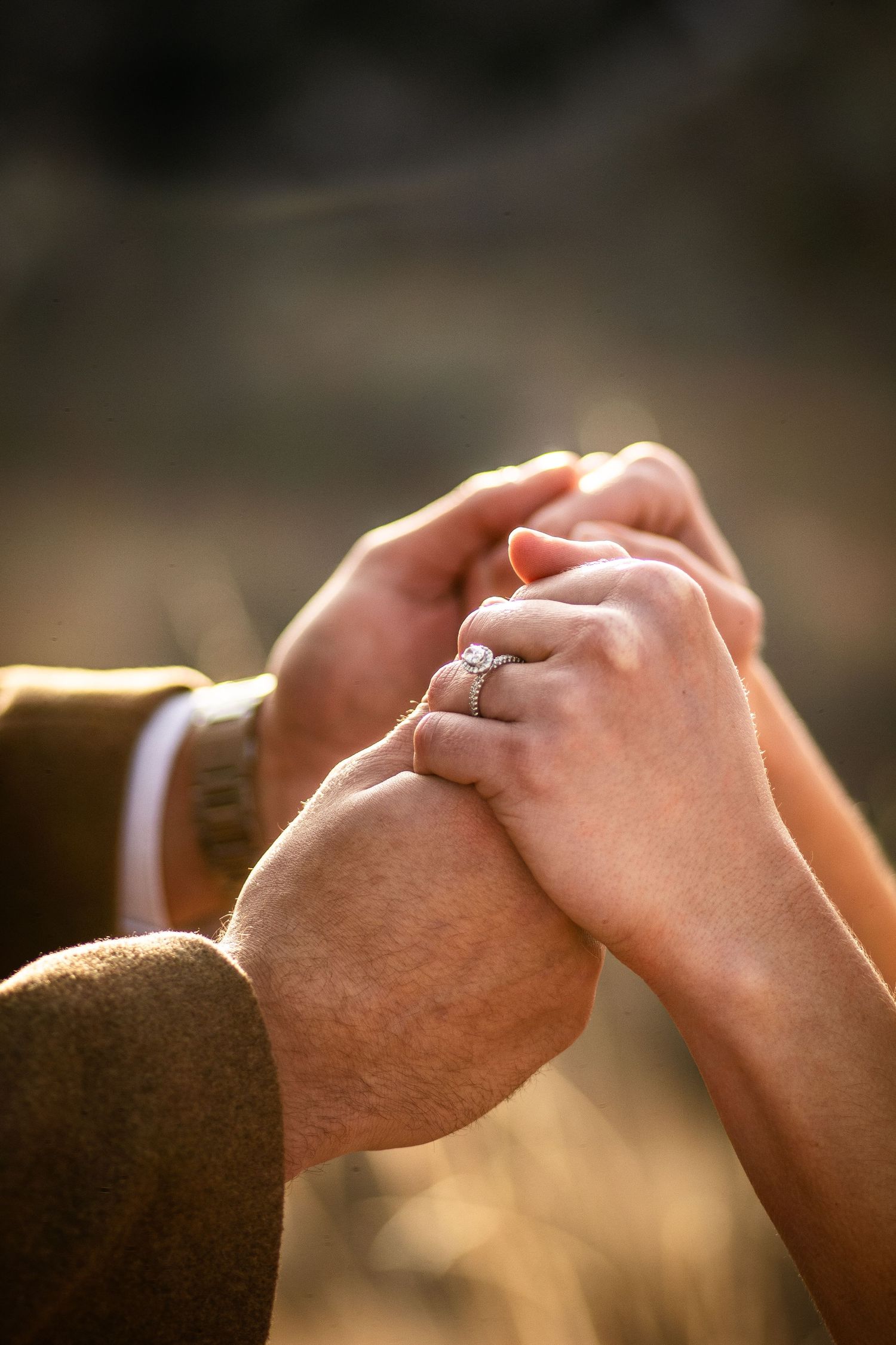 Close-up of couple holding hands showing diamond engagement ring in warm golden light.