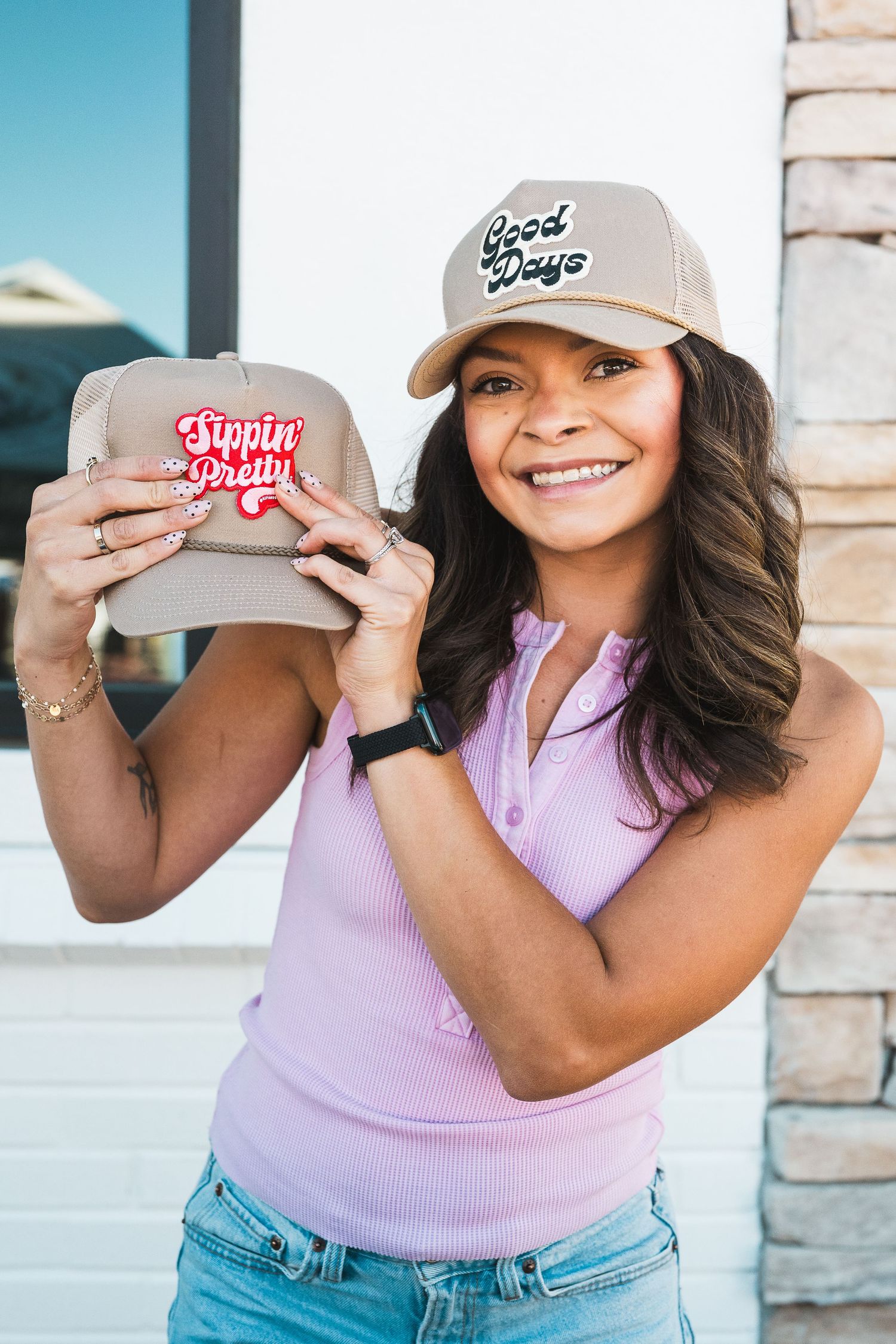 Smiling woman wearing a tan trucker hat holding a second tan cap with a Sippin Pretty patch outdoors.