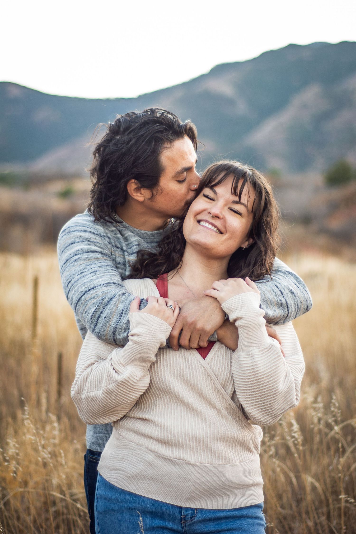 Man embracing and kissing woman on cheek in golden field with mountain background.