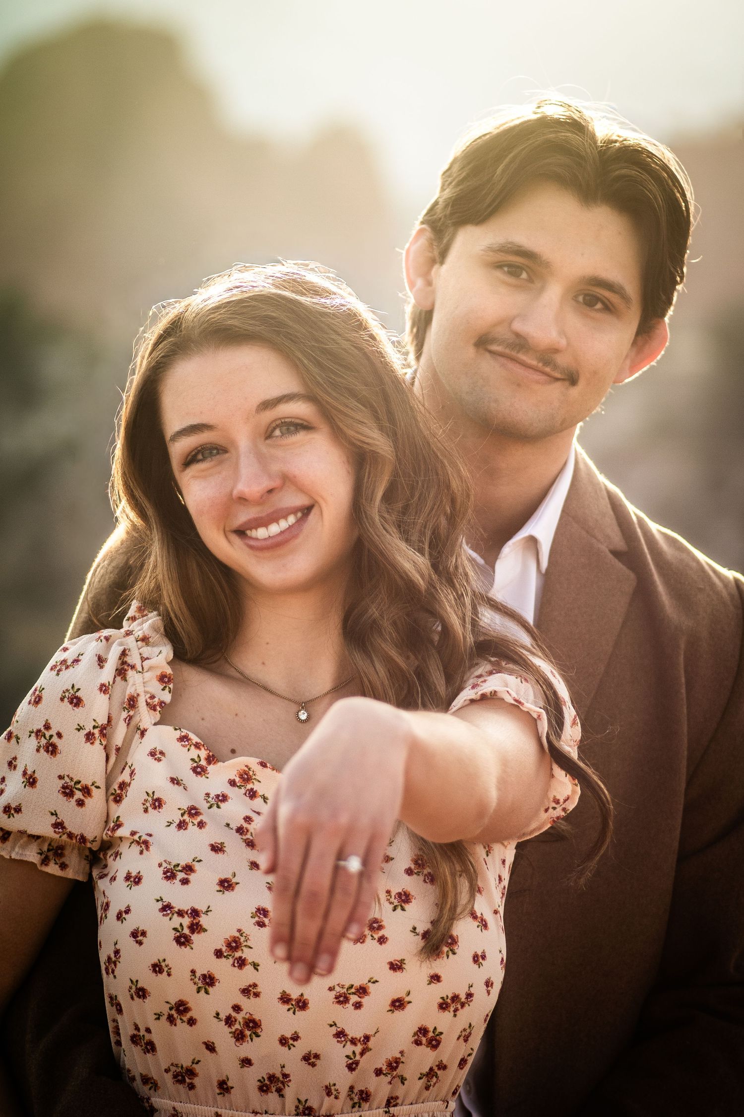 Smiling young couple posing together in warm golden sunset light with engagement ring visible.
