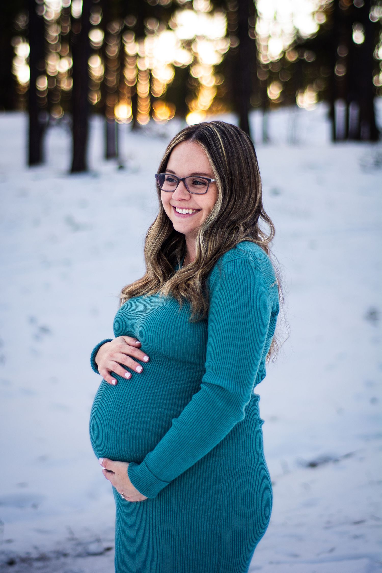 Smiling pregnant woman in teal dress holding baby bump in snowy winter forest at sunset.