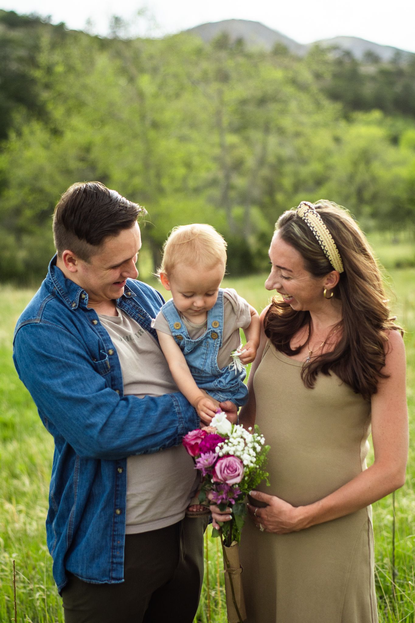 Happy family with dad holding baby and pregnant mom holding flowers in a green outdoor setting.