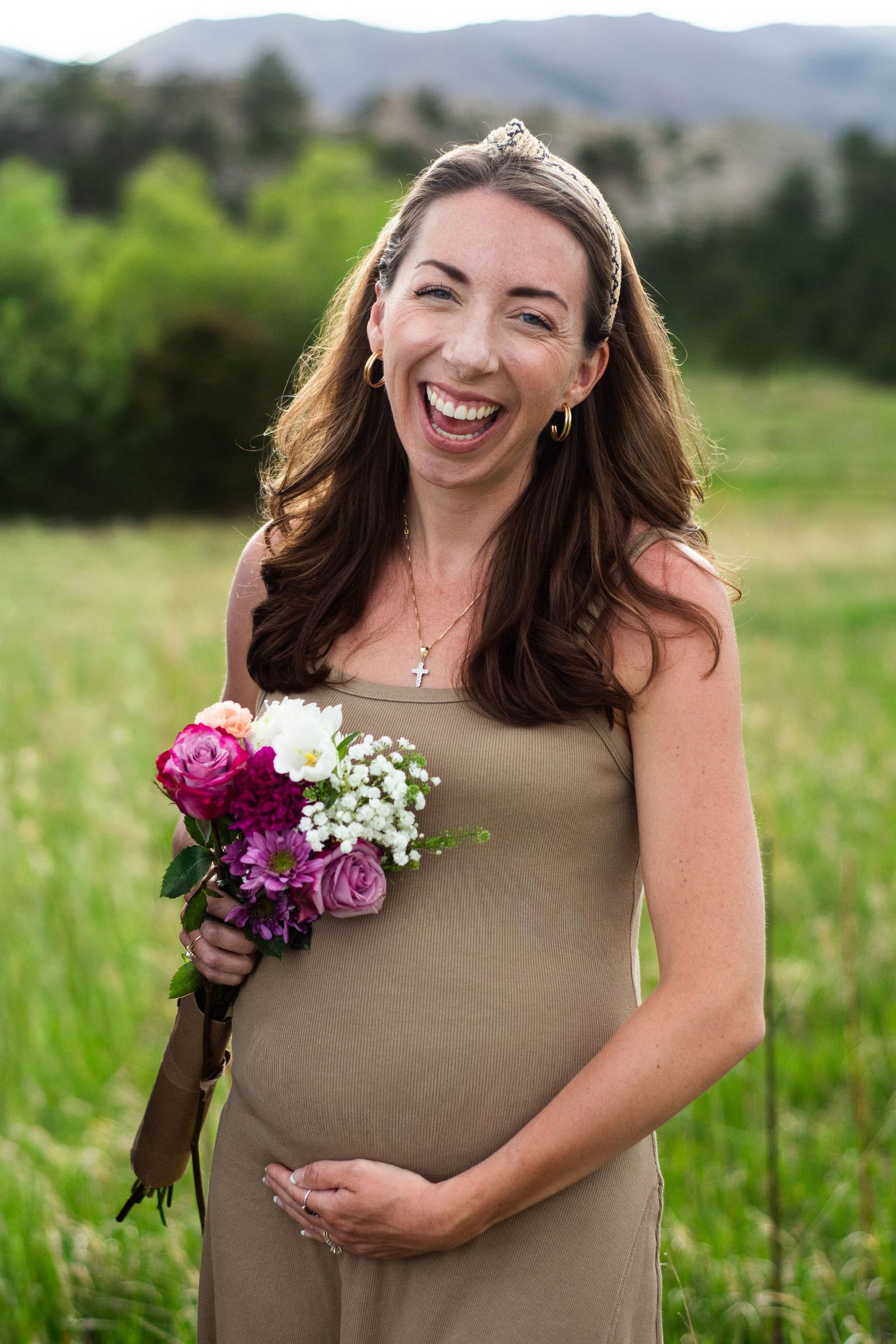 Smiling pregnant woman in tan dress holding pink and white flower bouquet in green meadow with mountains behind her.