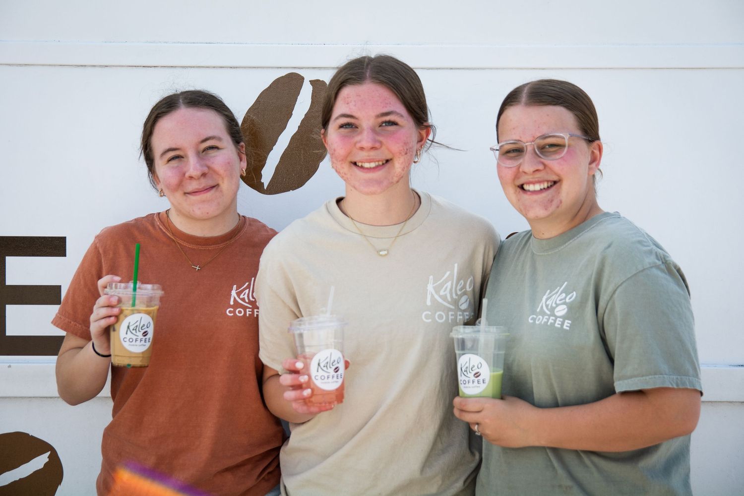 Three smiling women holding iced coffee drinks pose together outside a coffee shop wearing branded t-shirts.