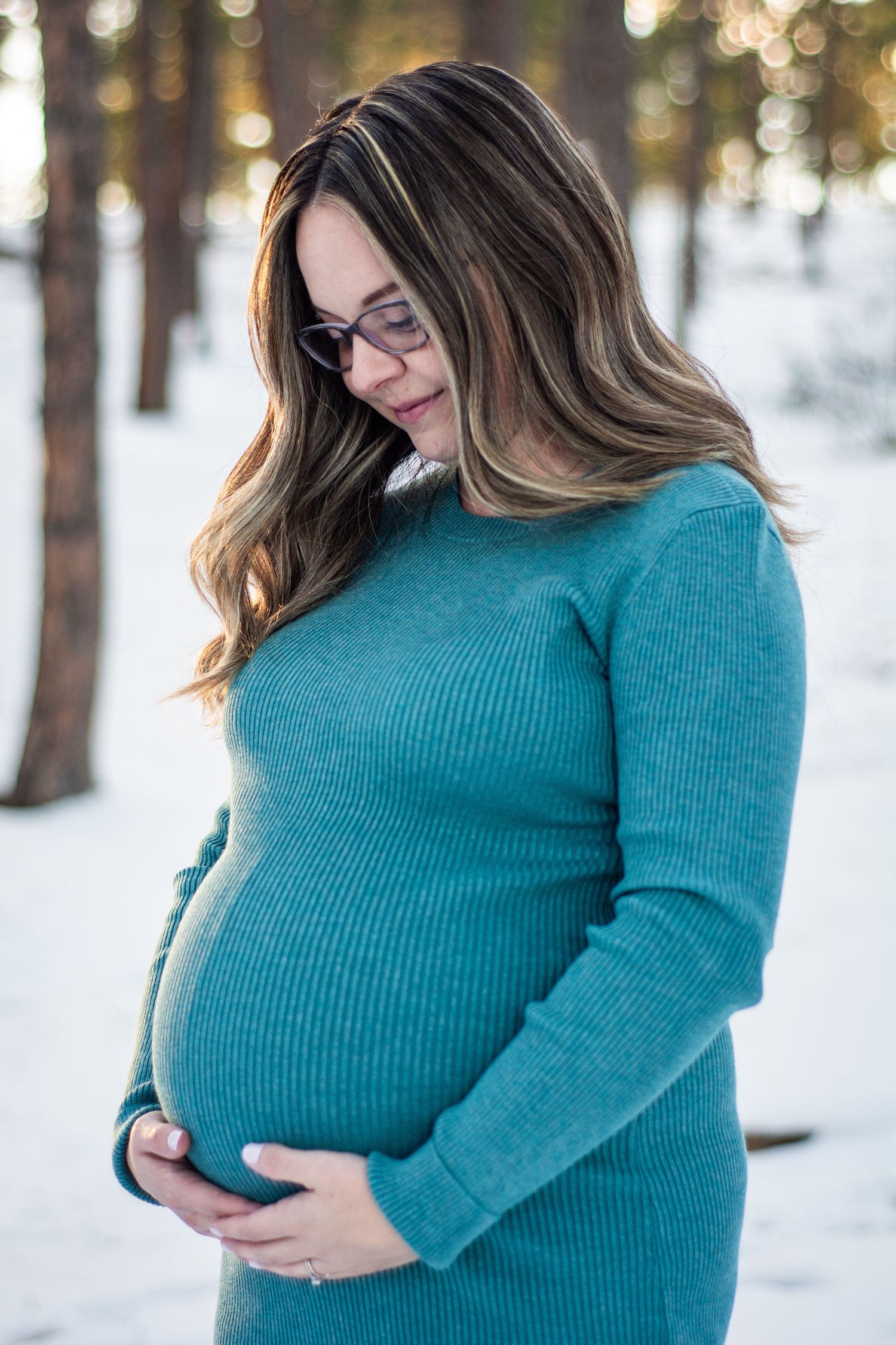 Pregnant woman in teal sweater cradling baby bump outdoors in snowy winter forest setting.