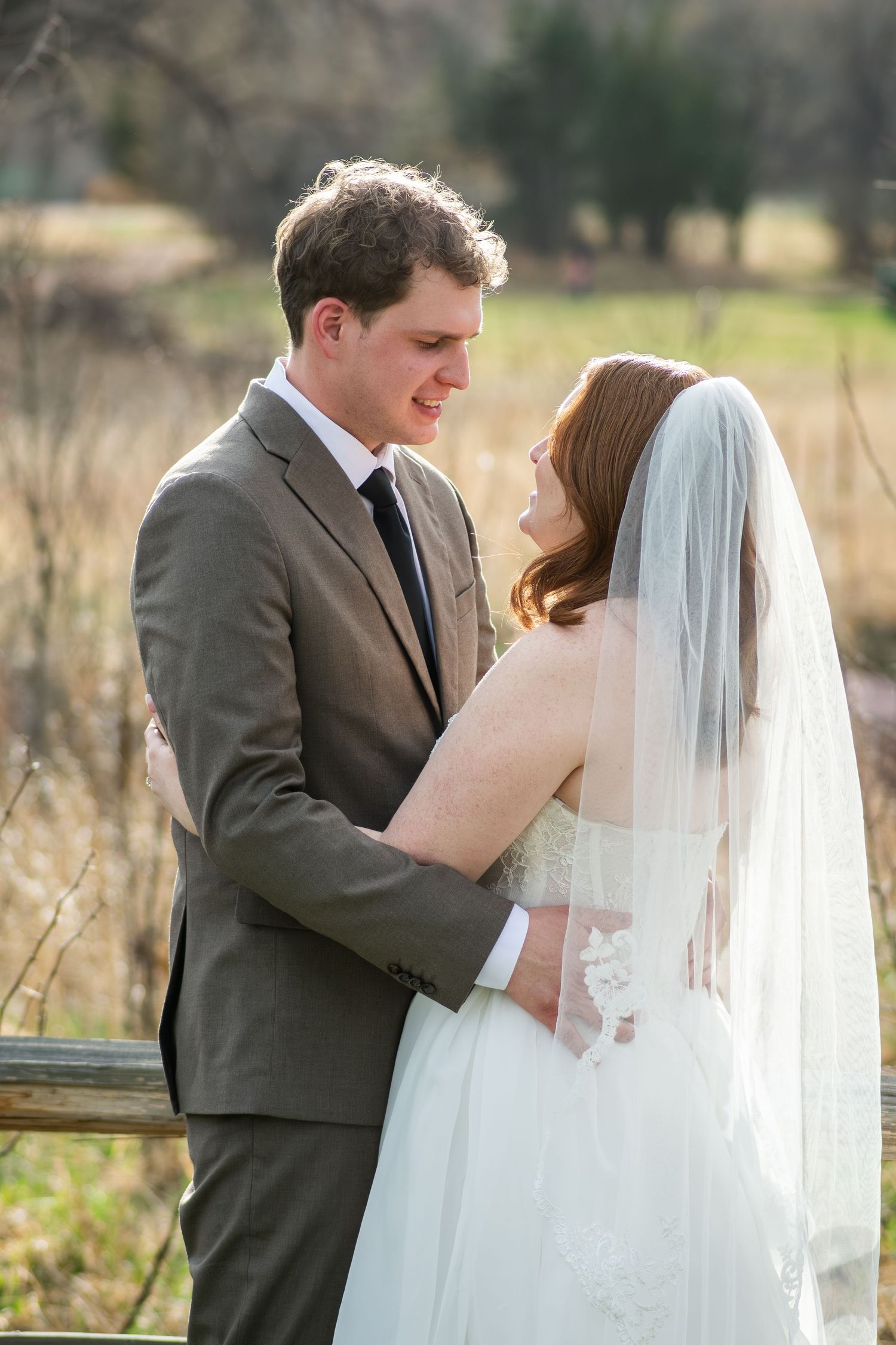 A bride in a white gown and veil embraces her groom in a gray suit outdoors during a romantic wedding portrait.