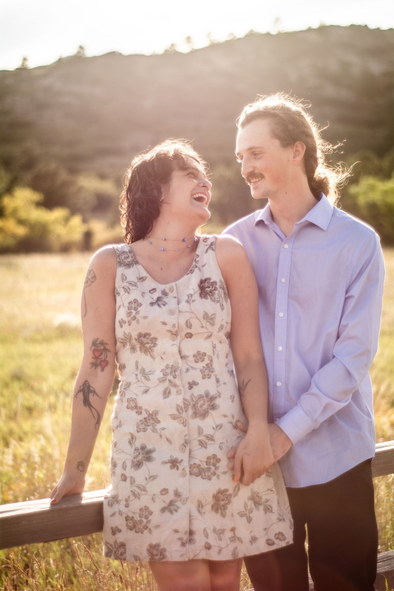 Laughing couple holding hands at golden hour in a sunny meadow, woman in floral dress with tattoos.