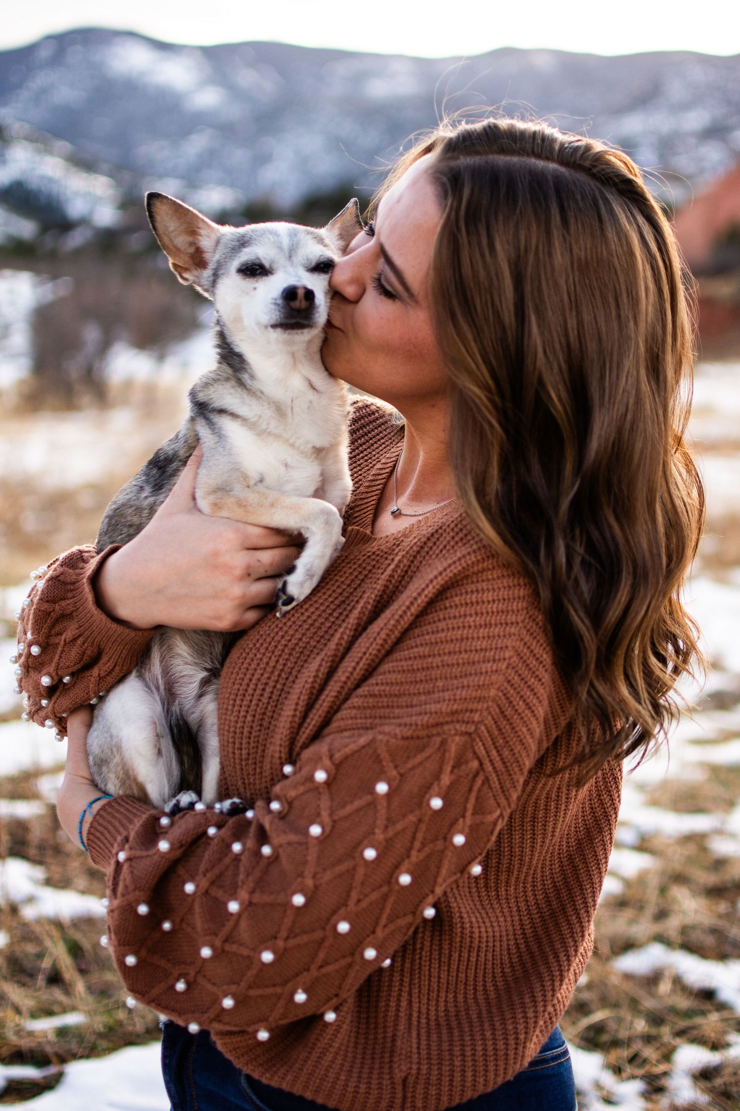 Young woman with wavy brown hair kissing her Chihuahua in snowy mountain setting.