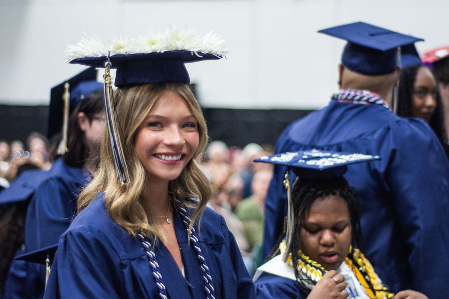 Blonde female graduate smiles in navy cap and gown during graduation ceremony with peers nearby.