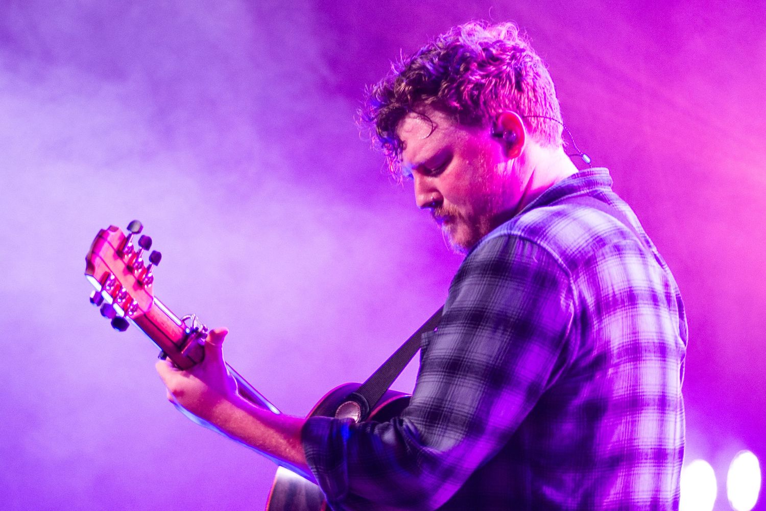 Musician playing acoustic guitar on stage under purple and pink stage lights with atmospheric fog.
