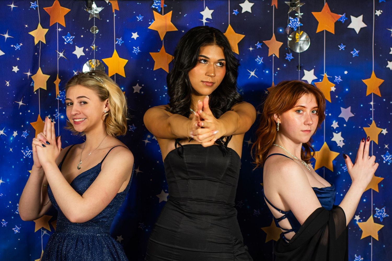 Three young women pose playfully in elegant dresses against a blue star-decorated prom backdrop.