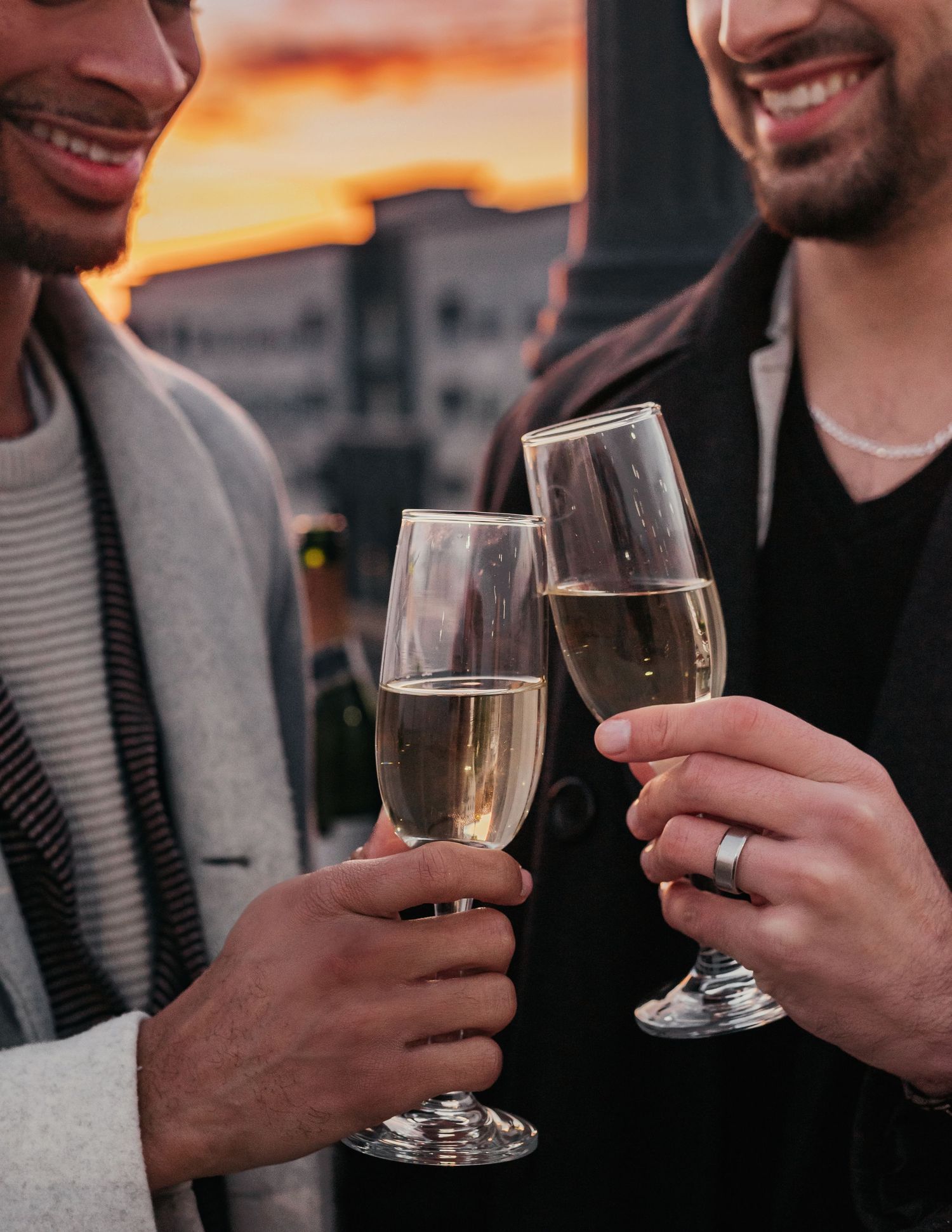 Two grooms clinking champagne glasses during golden hour rooftop engagement session at Bridgestreet
