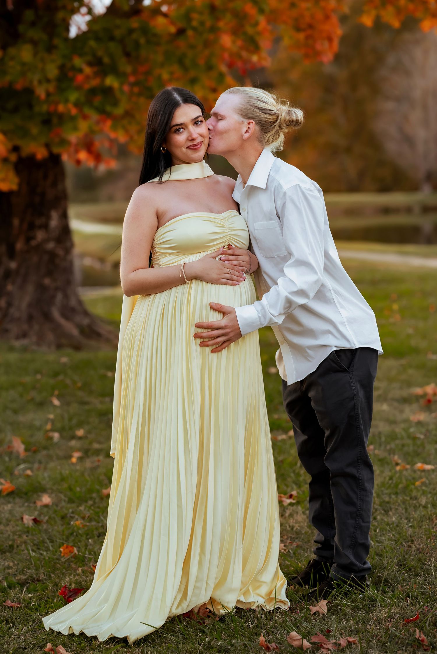Fall maternity session: woman in yellow gown smiling at camera, partner kissing her cheek, both holding belly, lake and trees blurred
