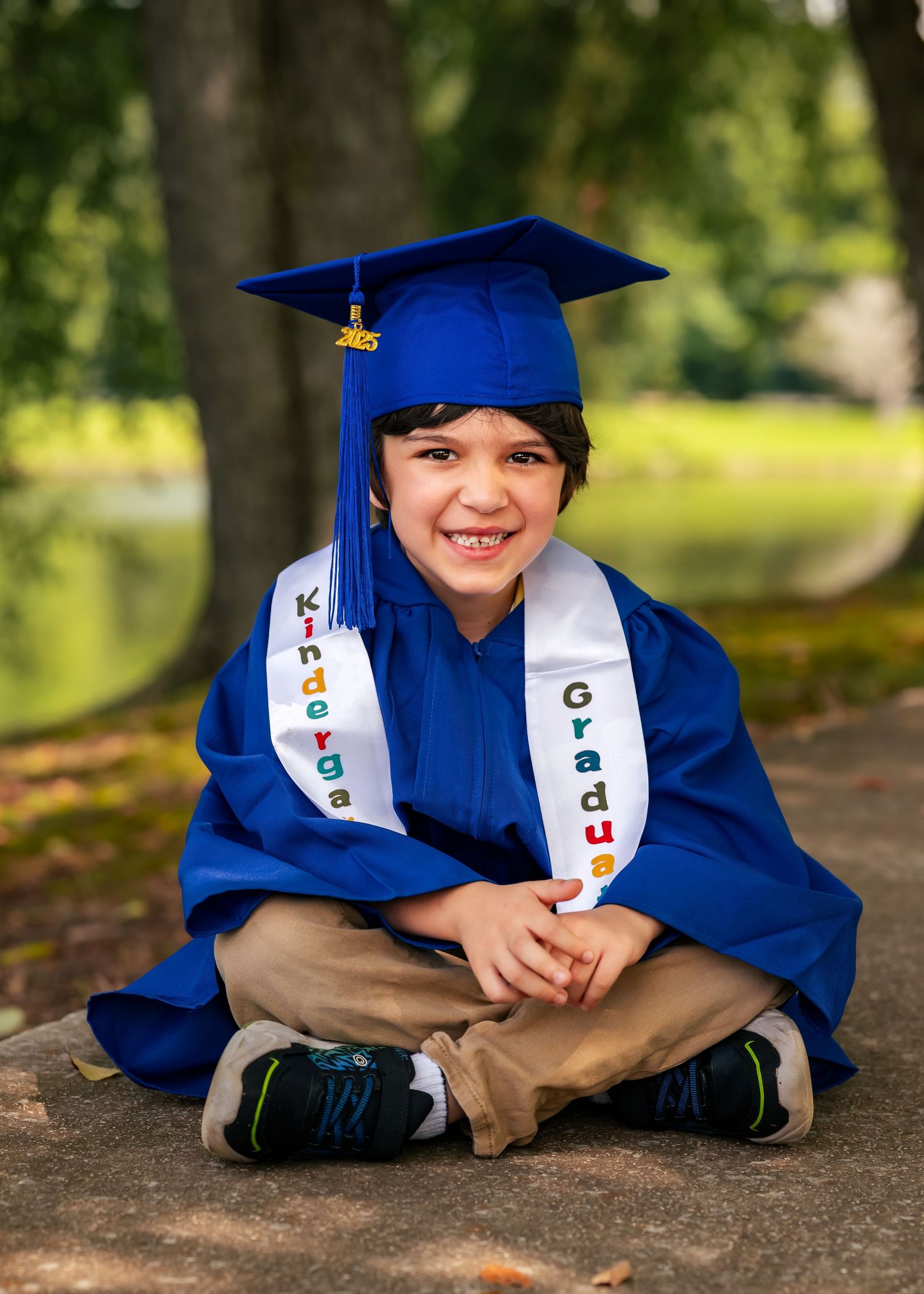 Portrait of smiling little boy who is a kindergarten grad wearing blue cap and gown next to Lady Ann Lake in Huntsville, AL.