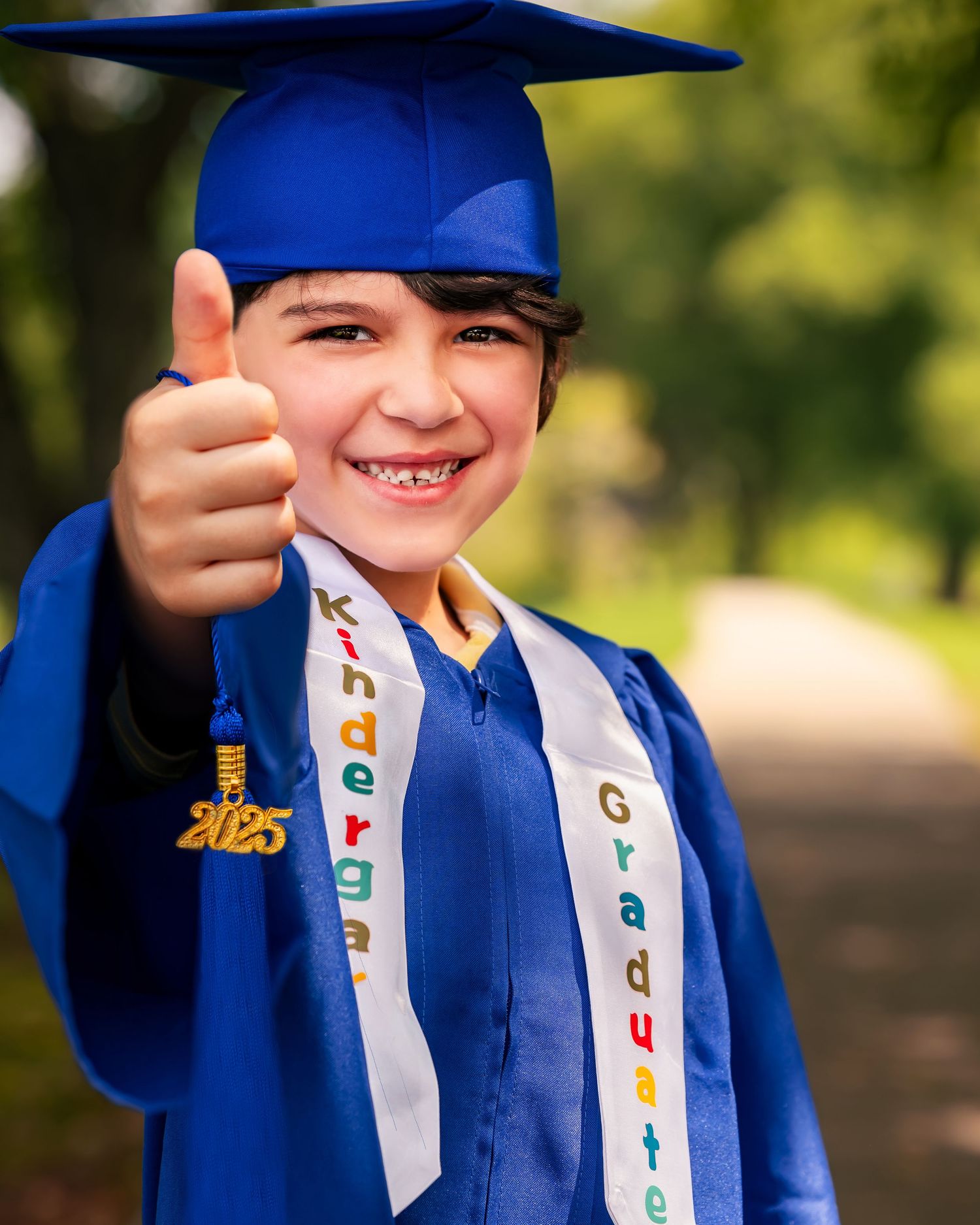 Kindergarten grad photo of little boy smiling, wearing blue cap and gown with a thumb up to the camera in Huntsville, Alabama.